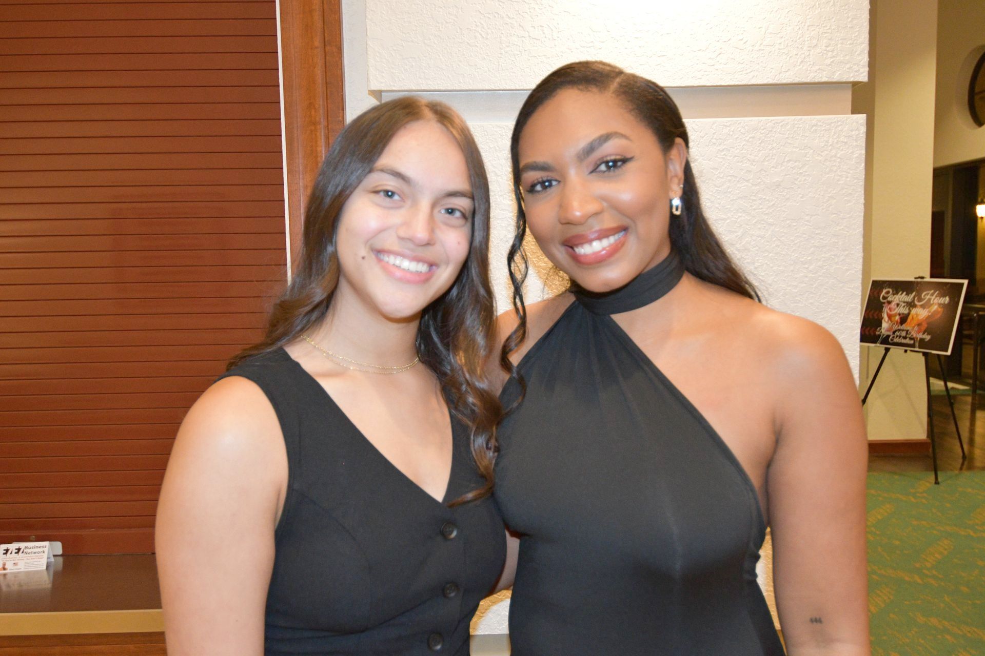 Two smiling people pose for a photo. One in a black halter dress, the other in a black button-down dress.