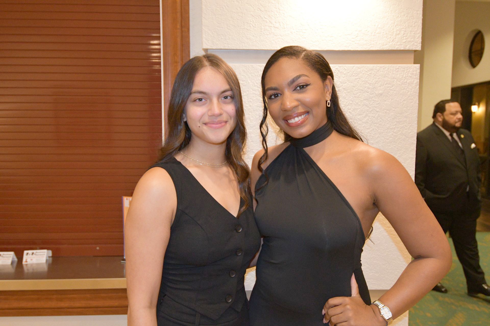 Two women in black dresses pose indoors, smiling, near a wall and a person in a suit.