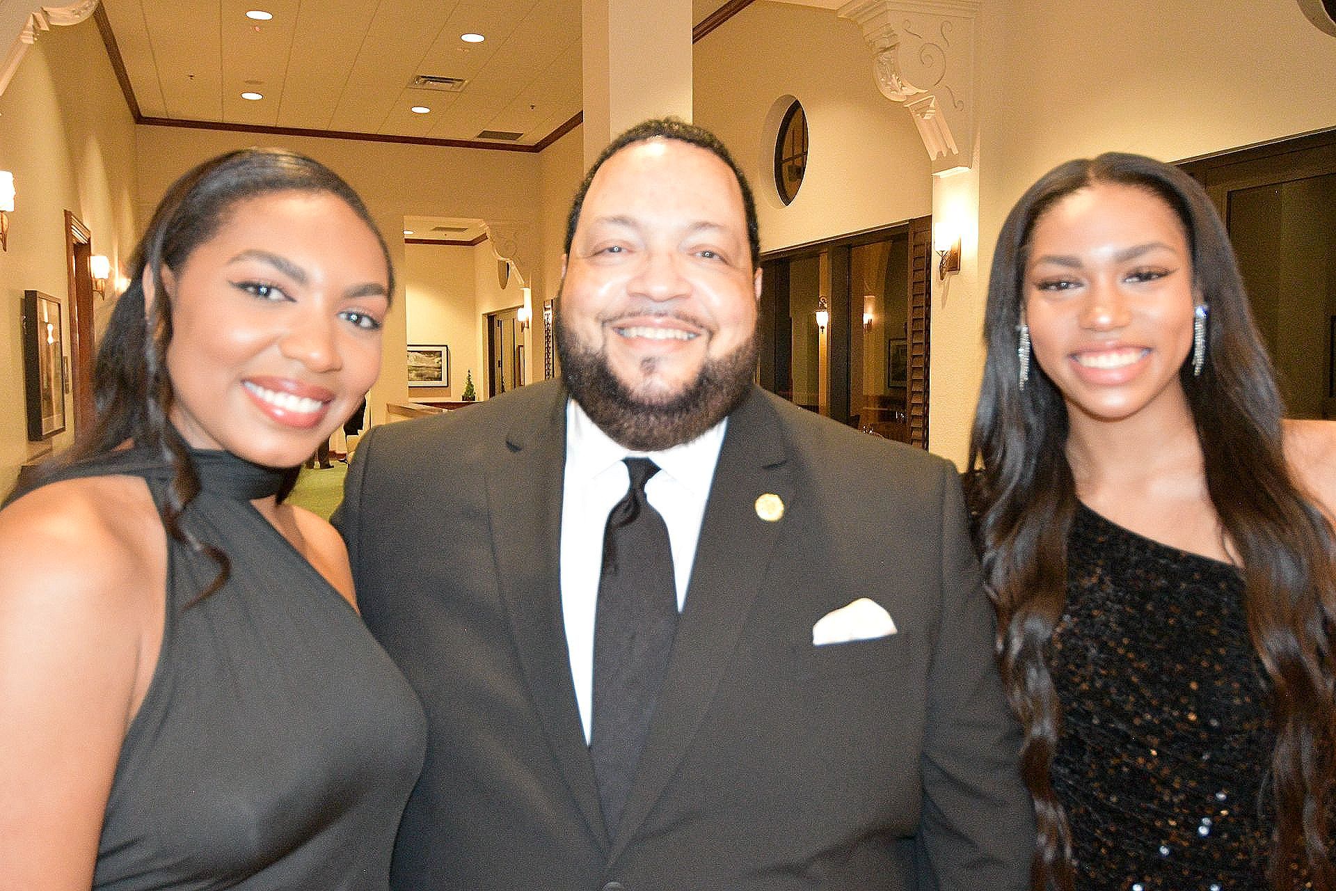 Three people smiling, posed in a hallway. Man in suit, two women in formal black dresses.