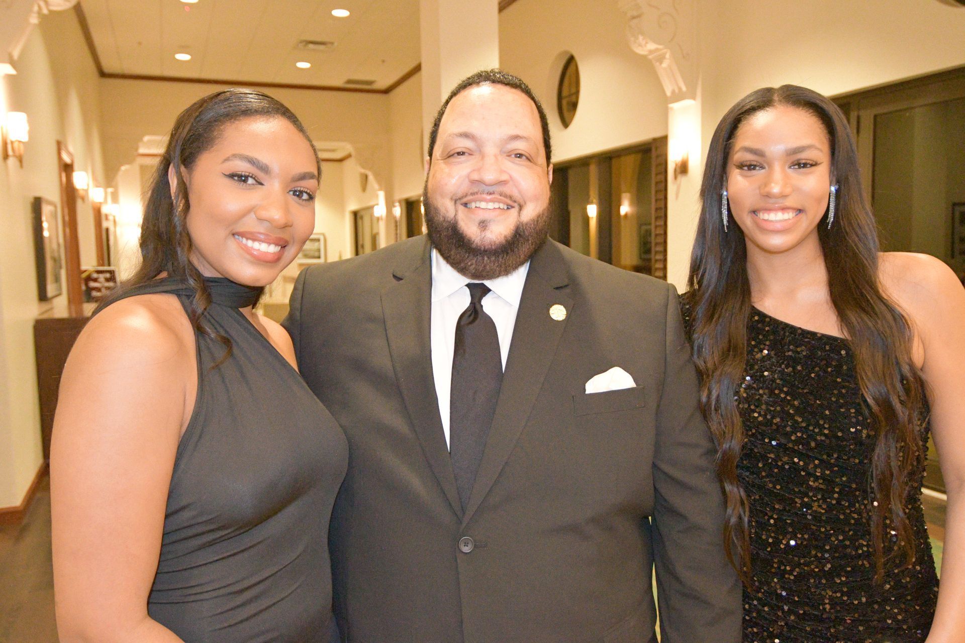Three people pose for a photo in a well-lit hallway. Man in suit smiles between two women in formal dresses.