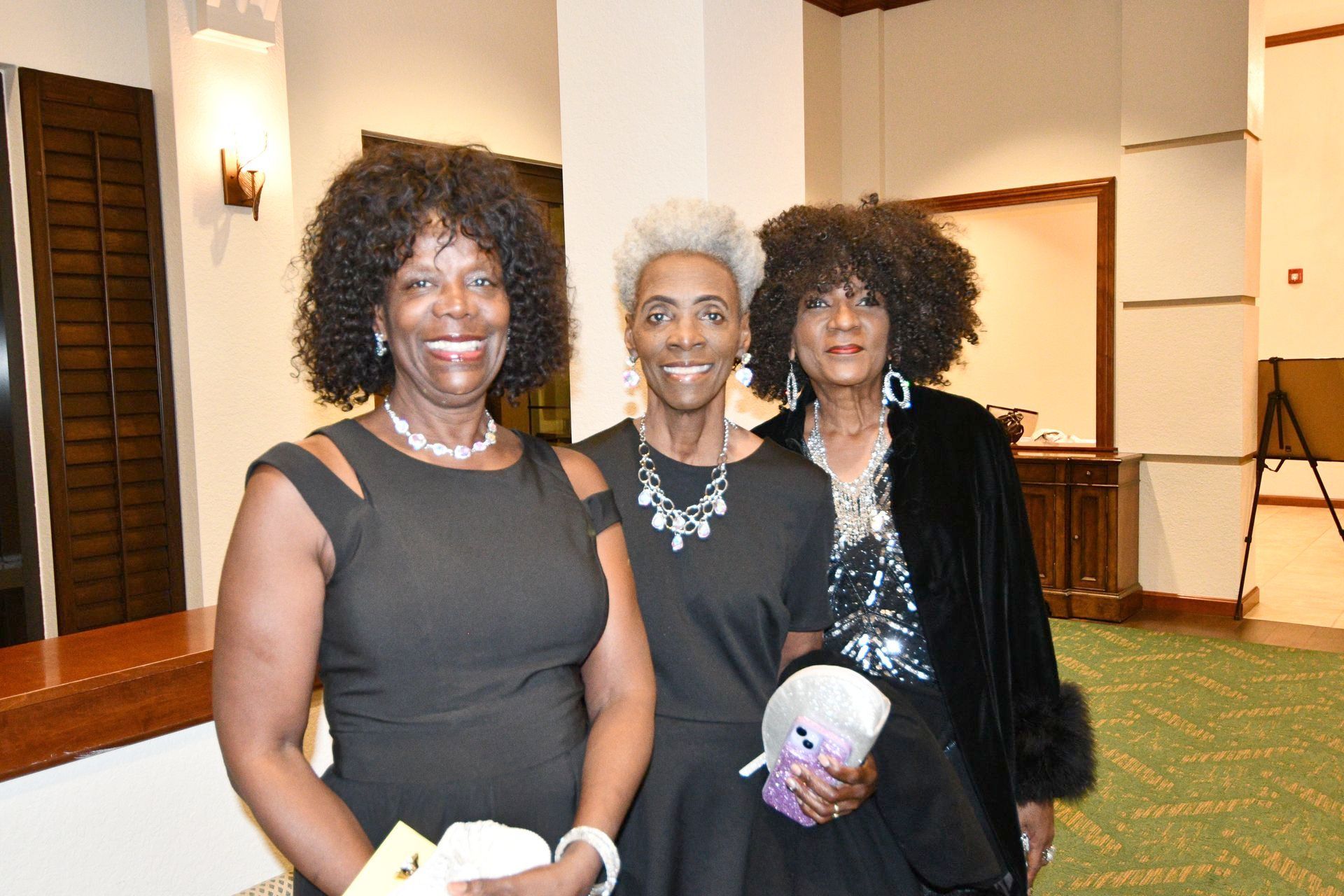 Three women smiling, posing for a photo. One in a black dress, one in a black top with silver necklace, and one in a sequin top with a fur coat.