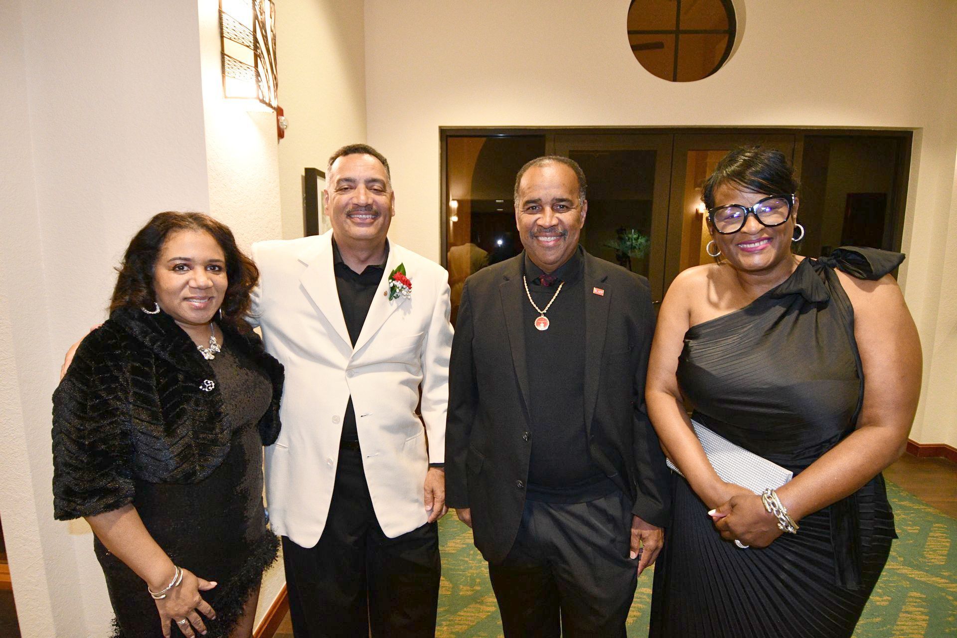 Four formally dressed people smiling indoors. Two men in suits, two women in dresses, possibly at an event.