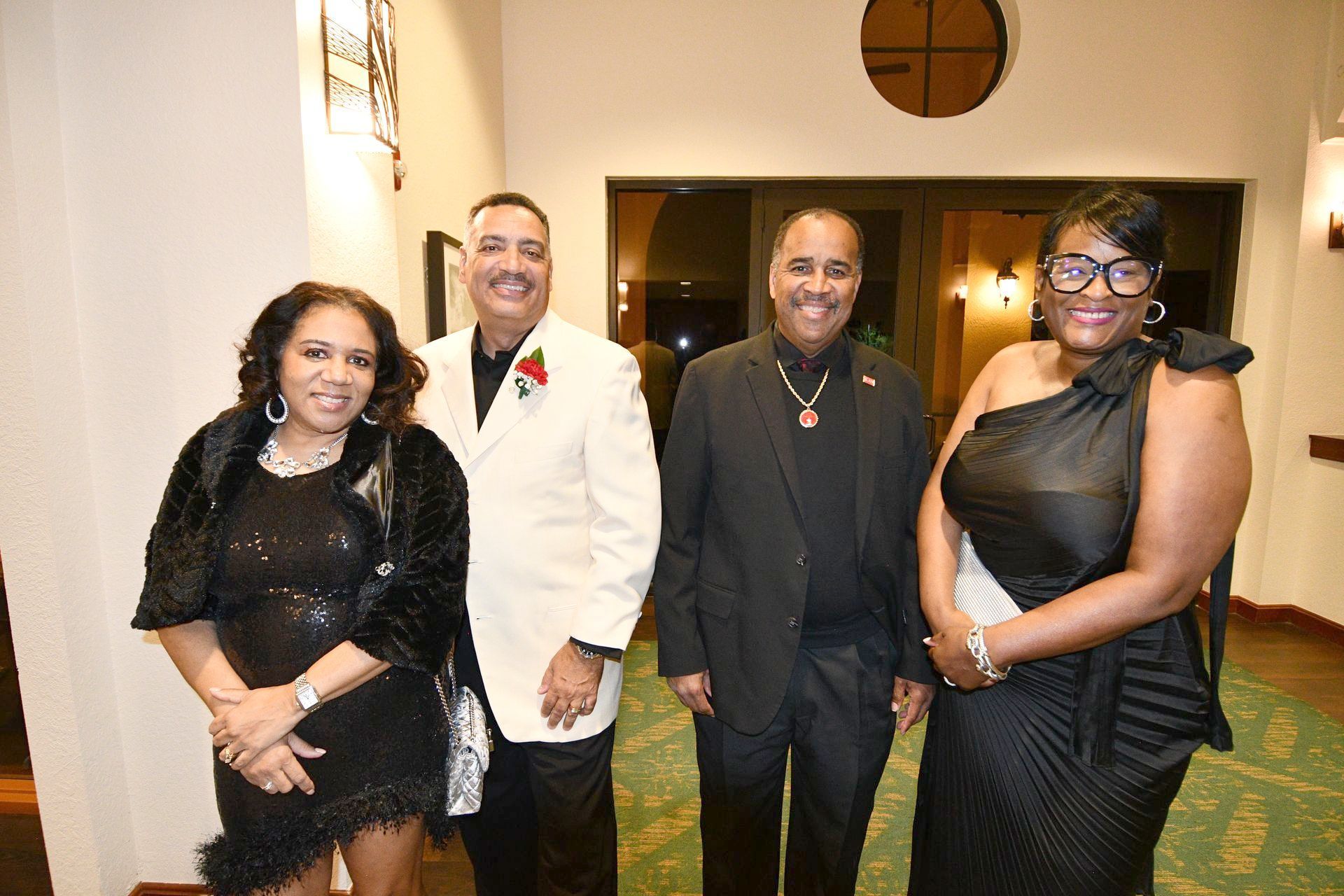 Four formally dressed people standing indoors; two men and two women smiling.