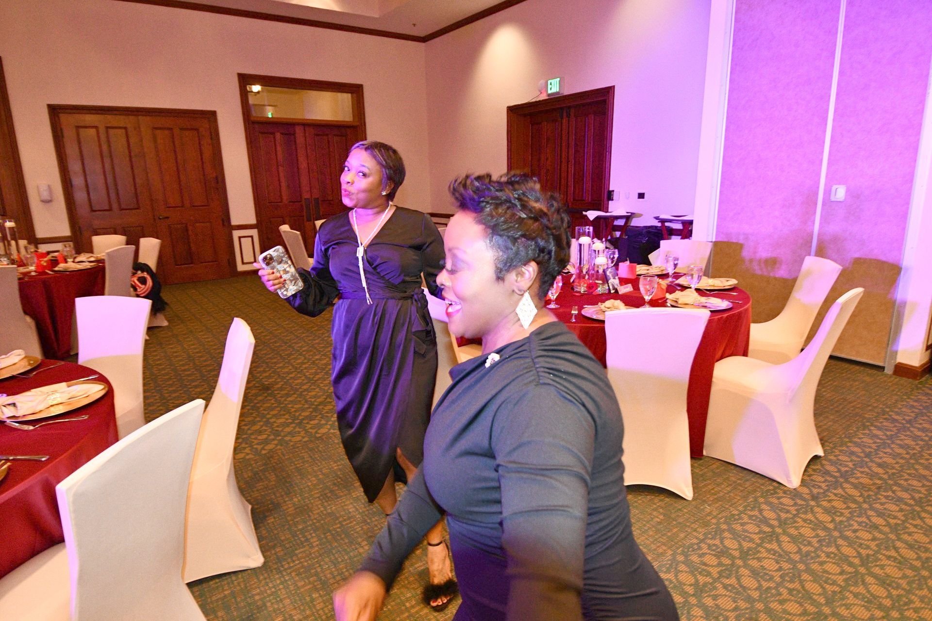 Two women in a banquet hall, one holding a phone, another smiling. Red tablecloths, white chairs. Purple and white lighting.
