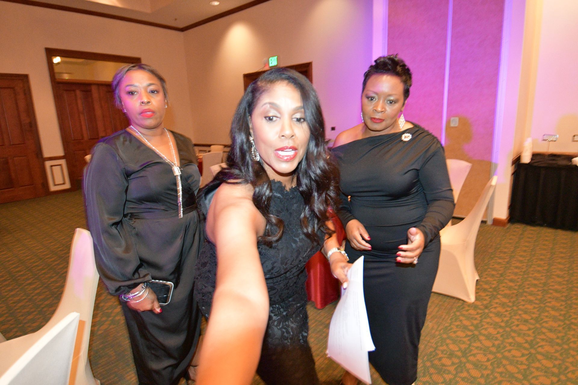 Three women in formal attire take a selfie indoors at an event.