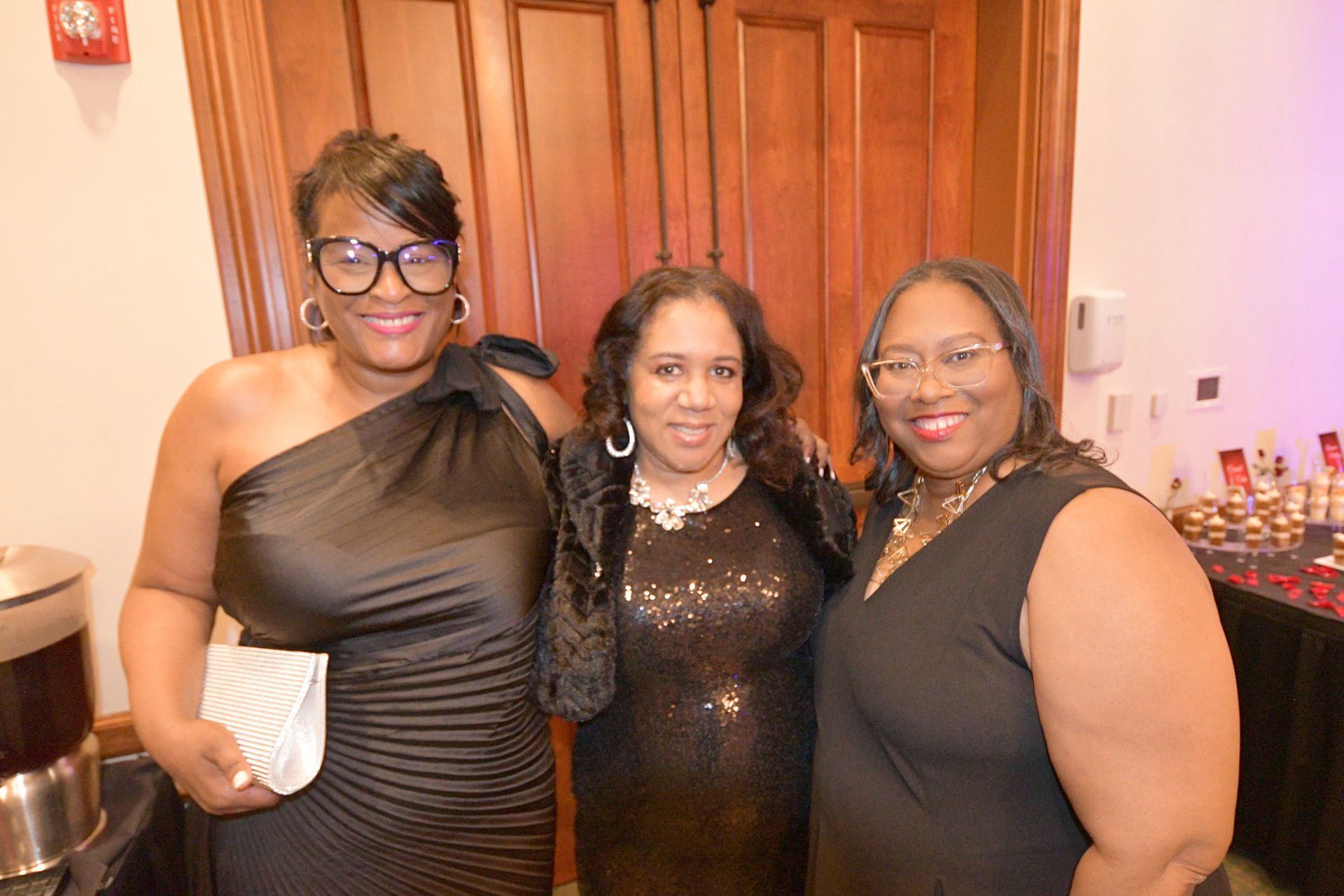 Three women in formal black attire pose together, smiling, at a social event.