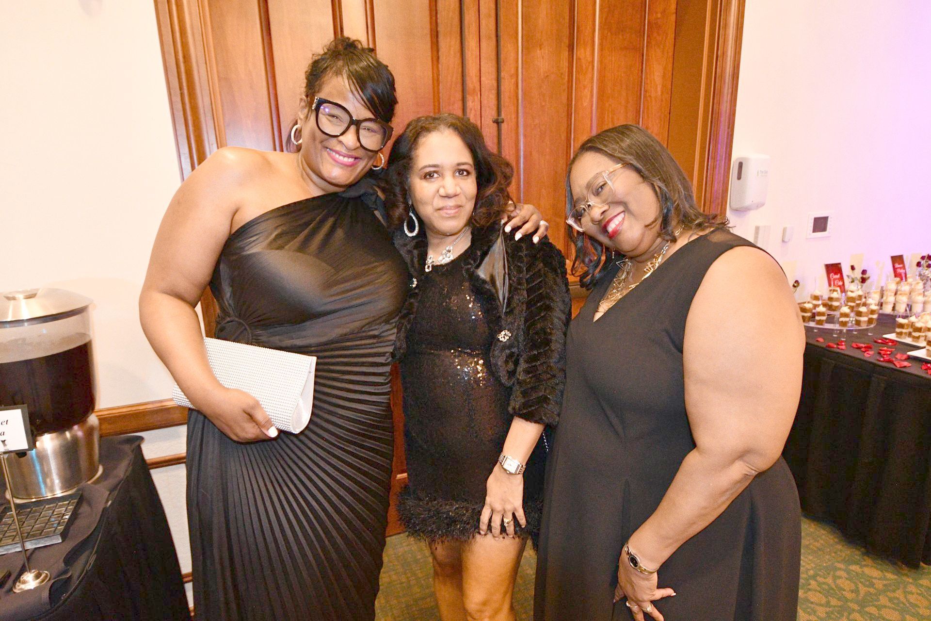 Three women in formal wear pose indoors, smiling. One in a black one-shoulder gown, two in dresses.