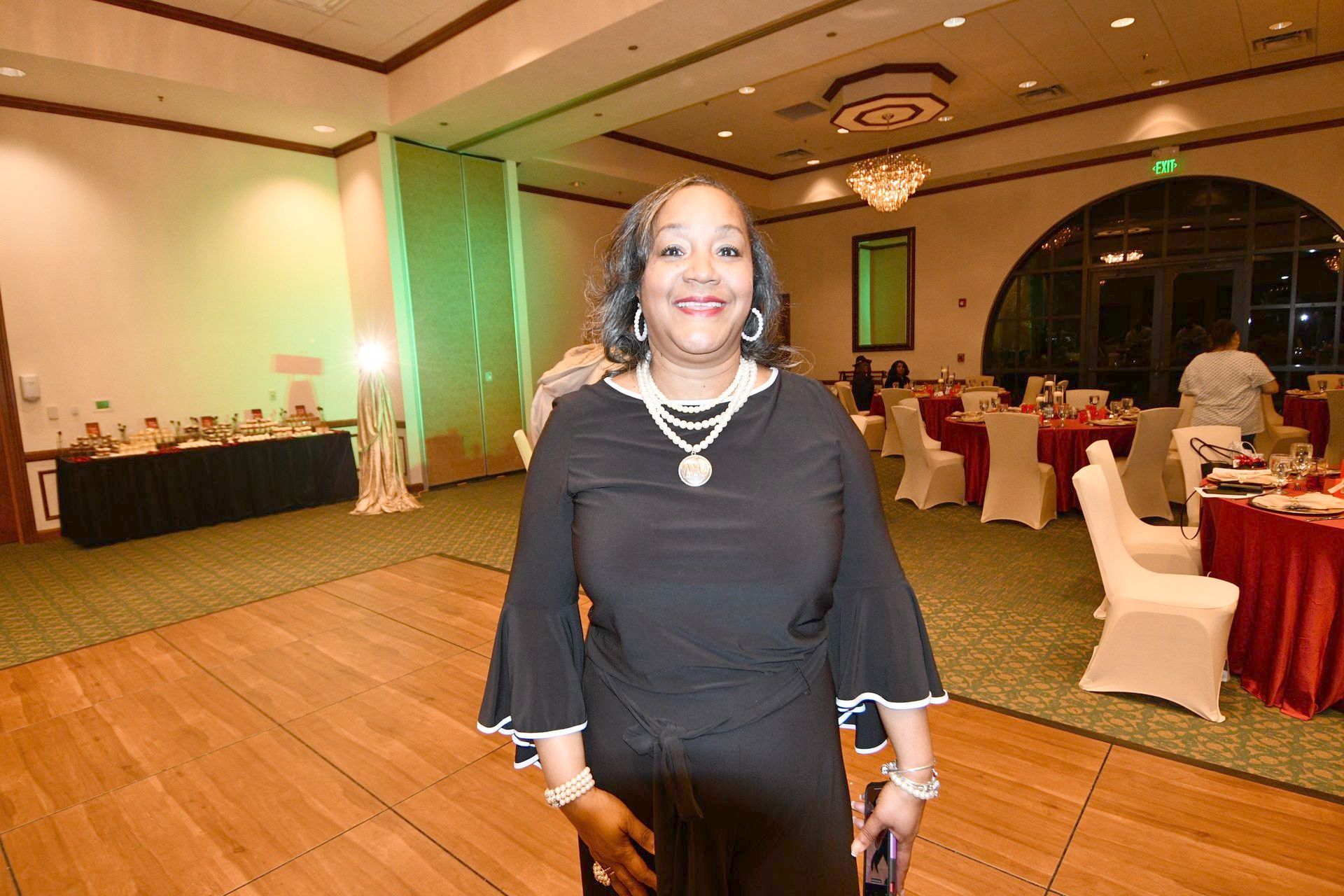Woman in black dress, smiles at camera, standing in a decorated ballroom.