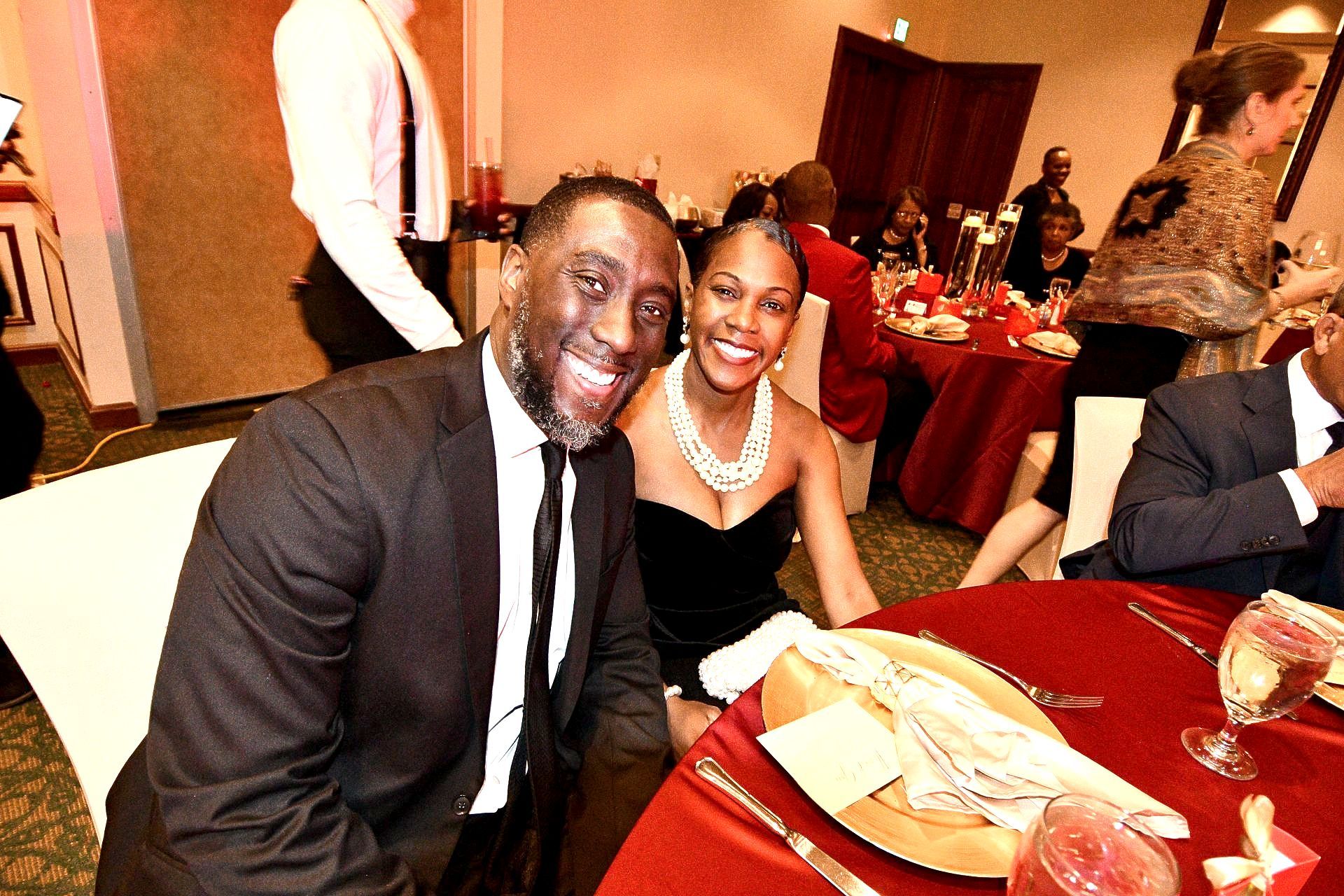 Man and woman smiling at a formal dinner table, red tablecloth, other guests blurred in the background.