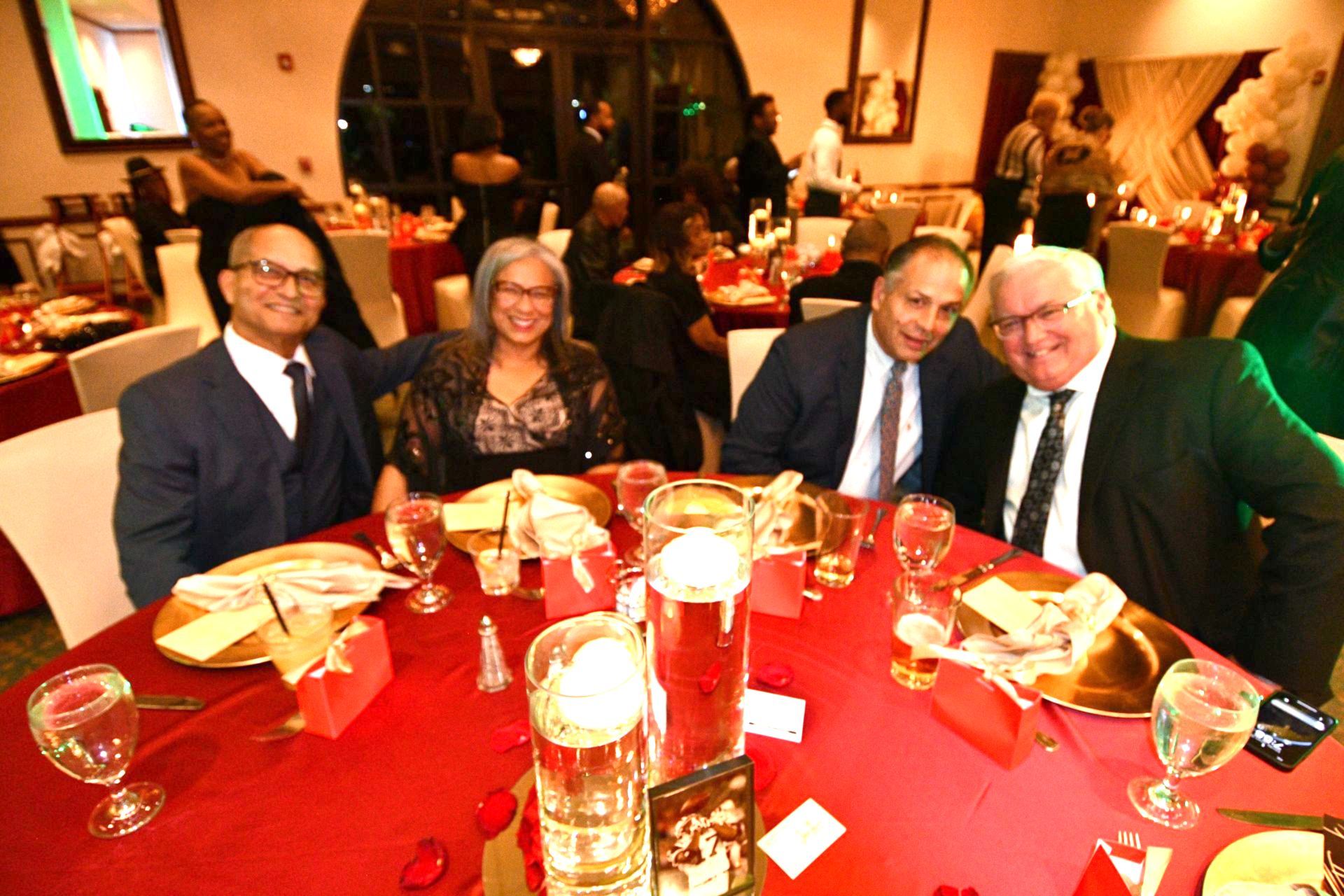 Four people at a round table, smiling. Festive red tablecloth, drinks, and candles.
