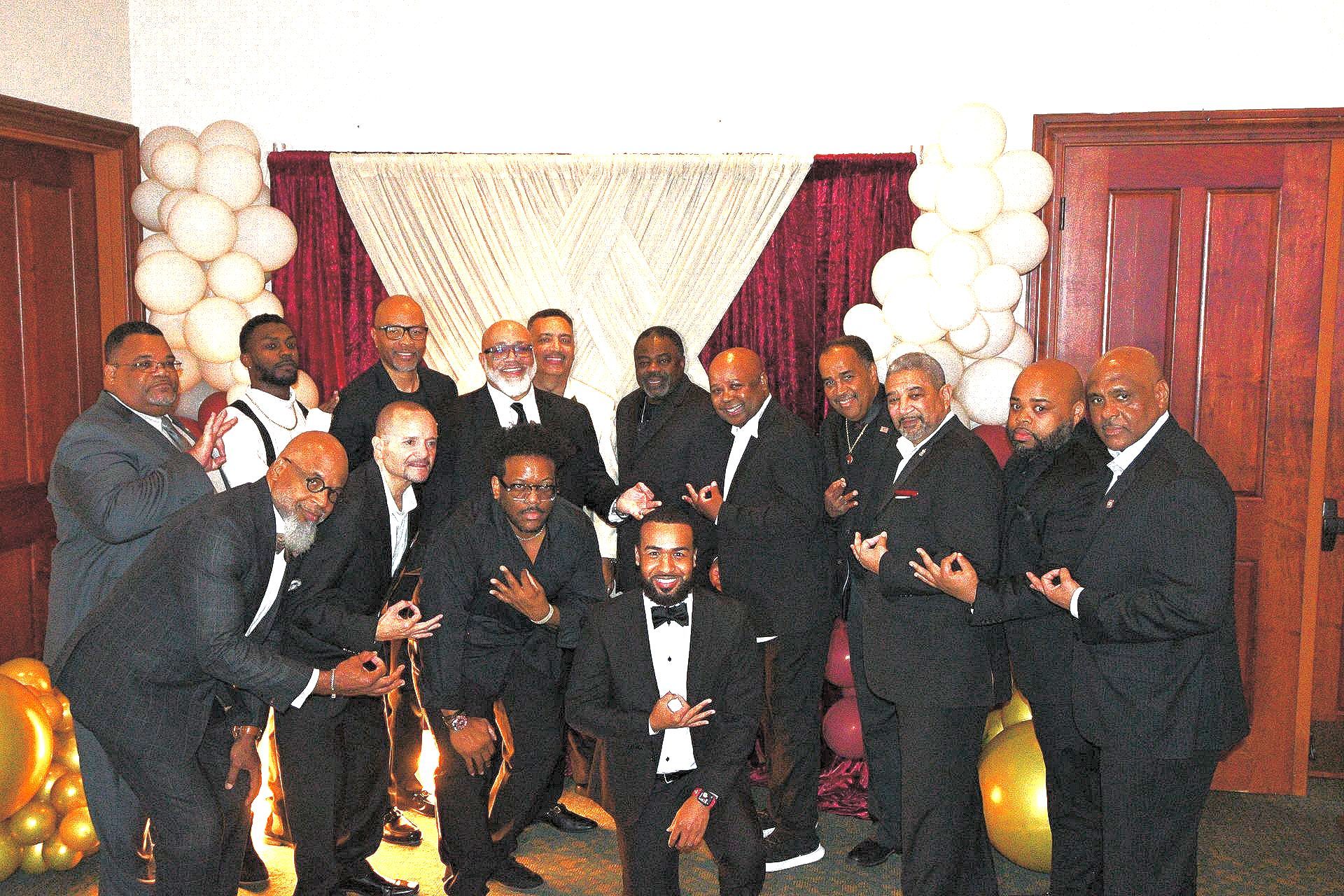 Group of men in suits posing together, some making a hand gesture, in front of a decorated background with balloons.