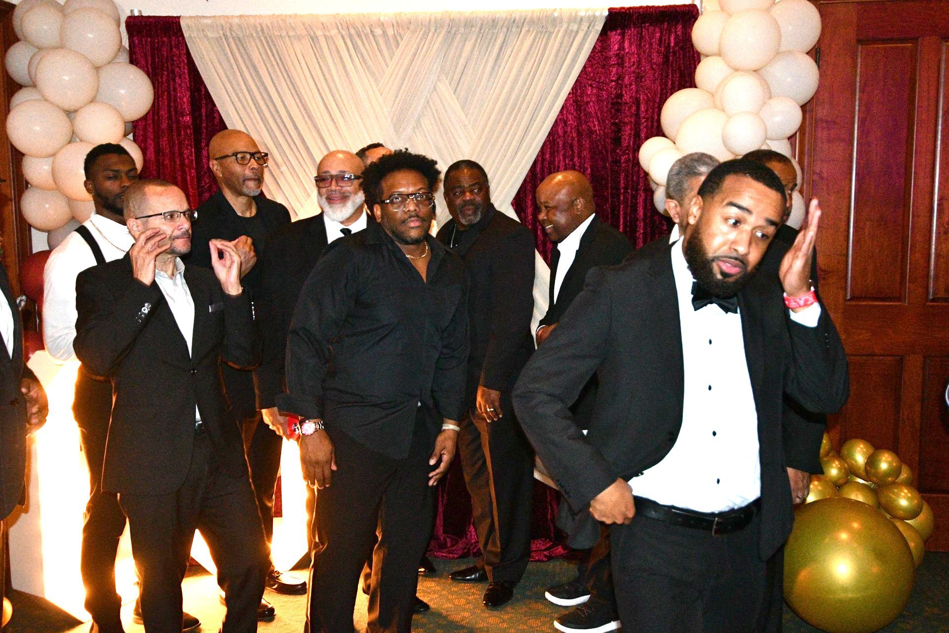 Group of men in formal attire, some with expressions of surprise, in front of a burgundy backdrop with balloons.