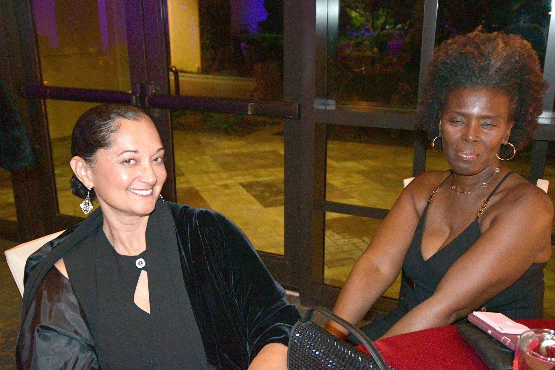 Two women at a table. One smiles, wearing black attire and a shawl. The other wears a dress, gazing away.
