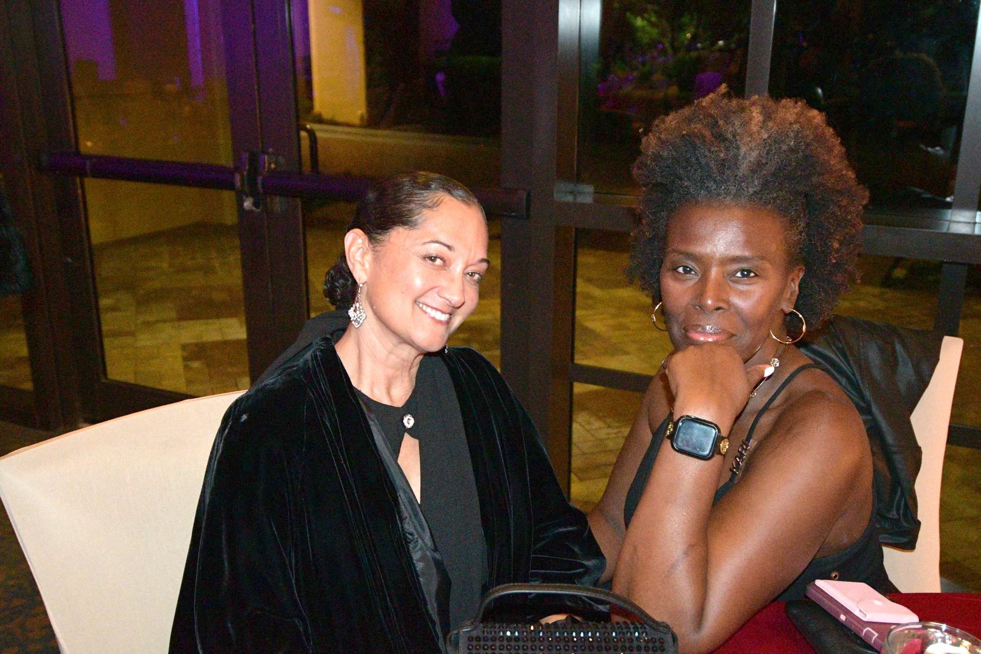 Two people, one in a black velvet jacket, the other with curly gray hair, seated indoors, smiling.