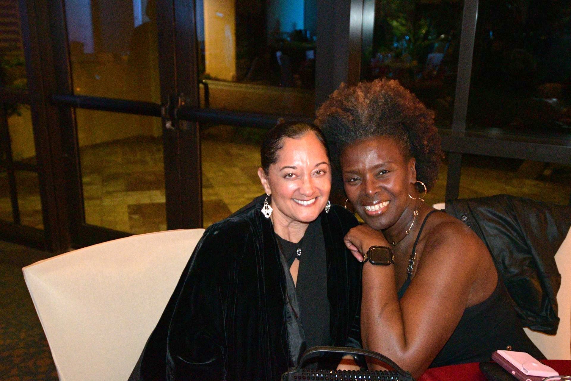 Two smiling women at an event; one in black velvet jacket, the other in a black dress, both with jewelry.