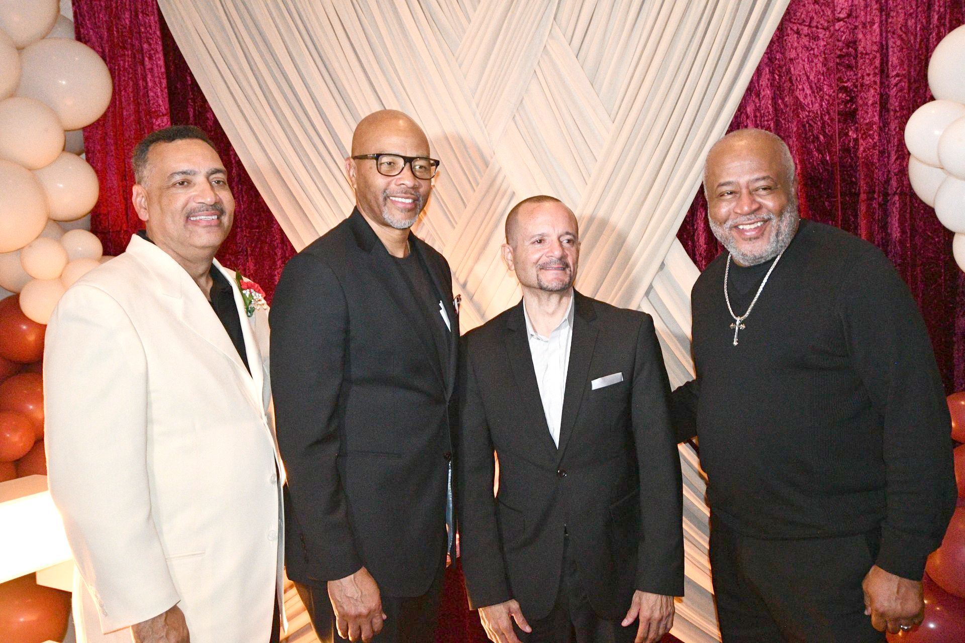 Four men in formal attire posing in front of a decorated backdrop with balloons.