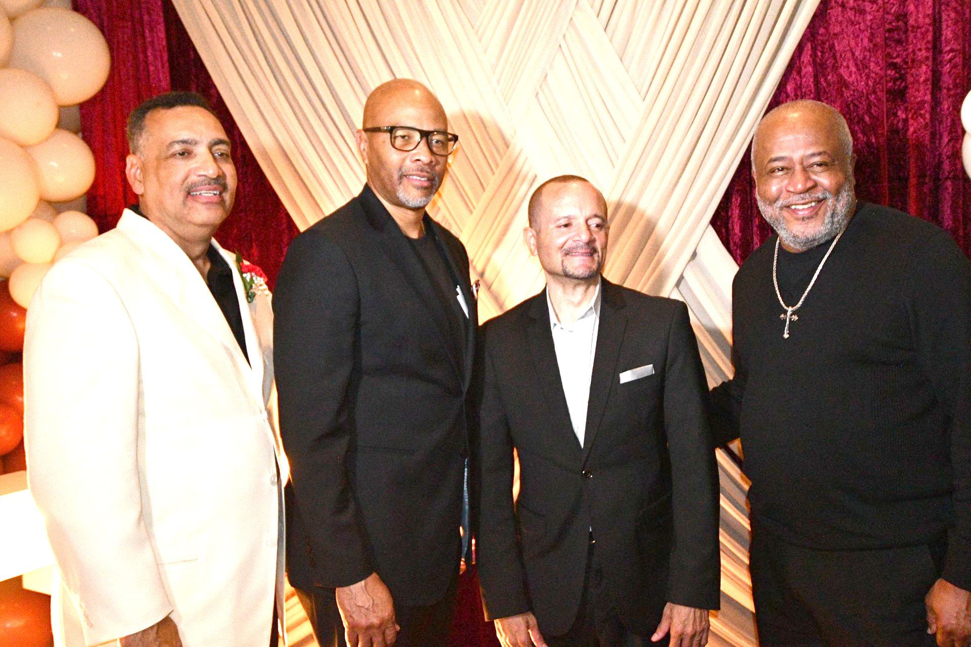 Four men in suits pose together in front of a draped backdrop with balloons.