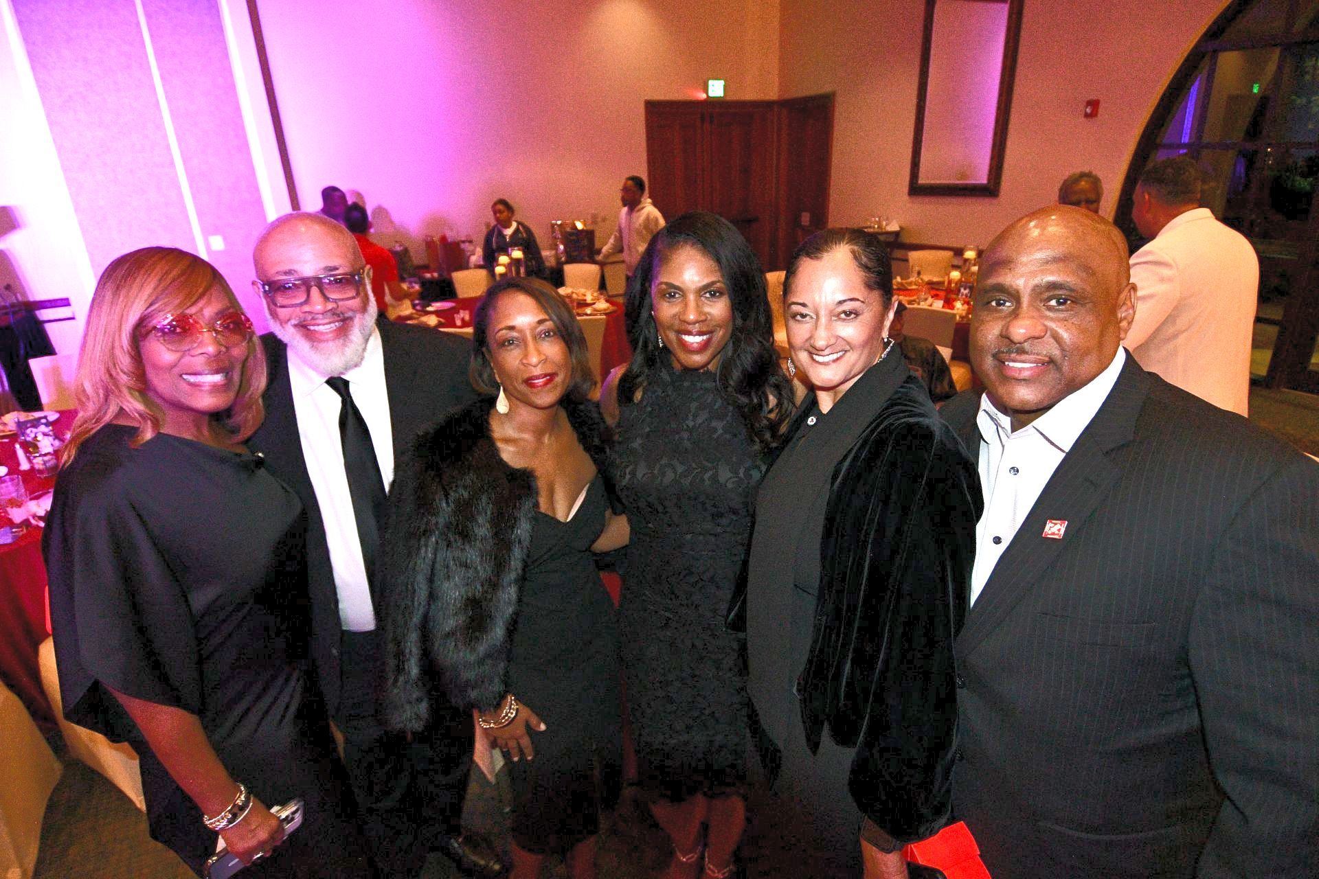 Group of six people posing at an event. They are in formal wear, inside a room with tables and lighting.