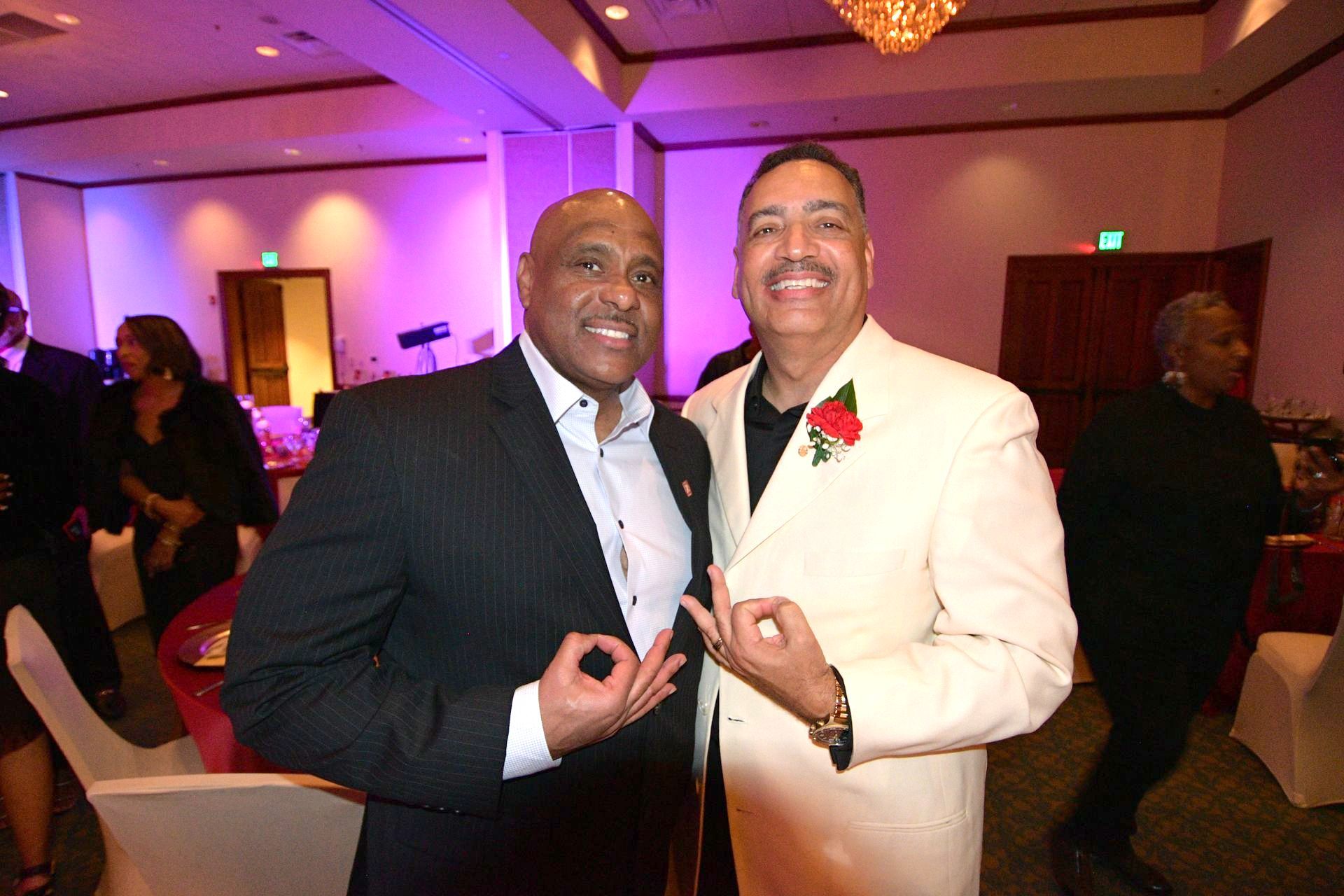 Two men in suits making okay hand gestures at a formal event, smiling.