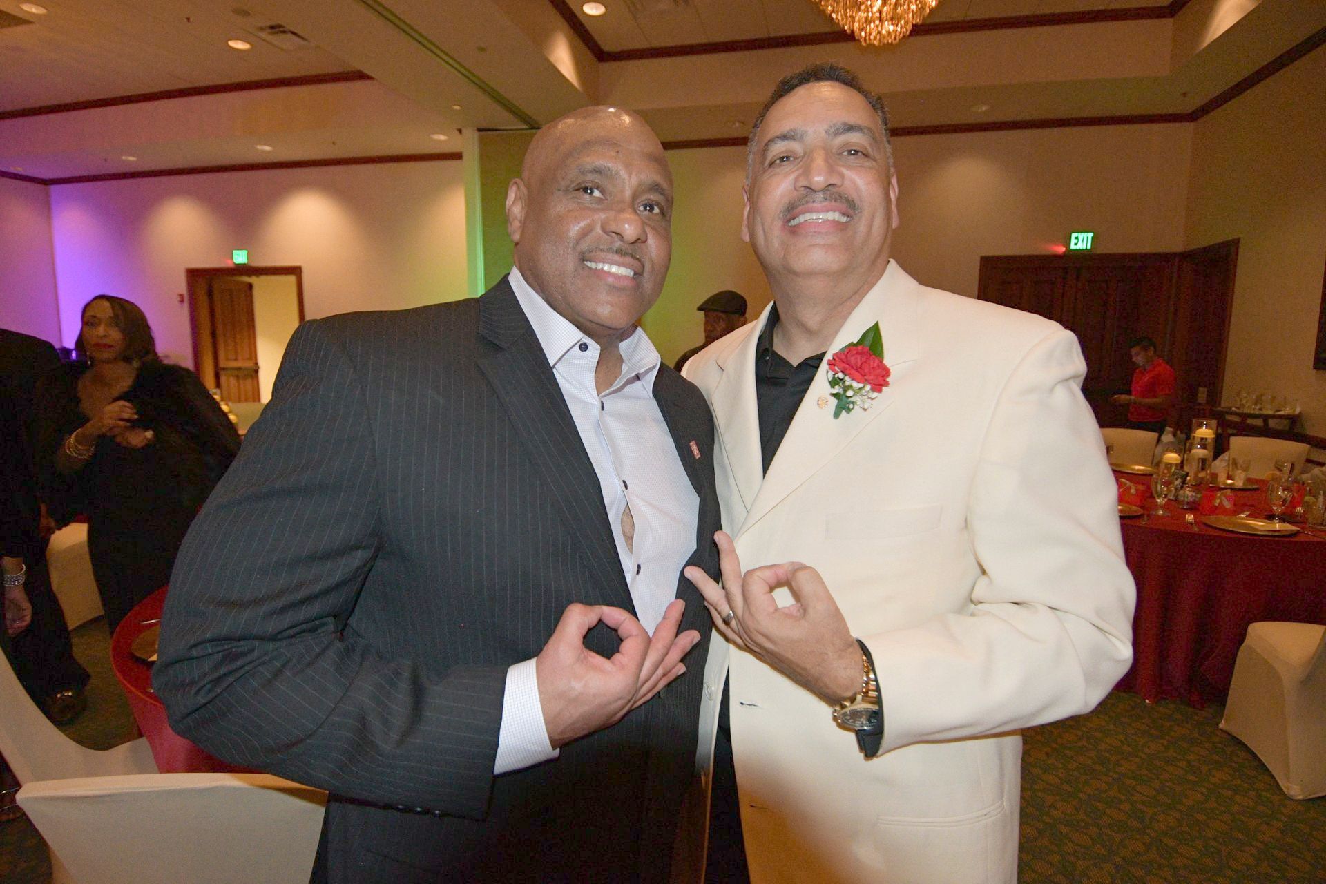 Two men in suits making hand gestures at an event with red tablecloths and decorations.