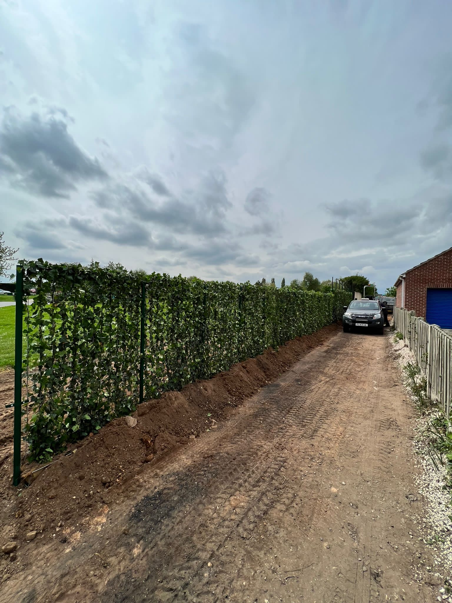 A car parked next to a green hedge, with dirt and gravel path on a cloudy day.