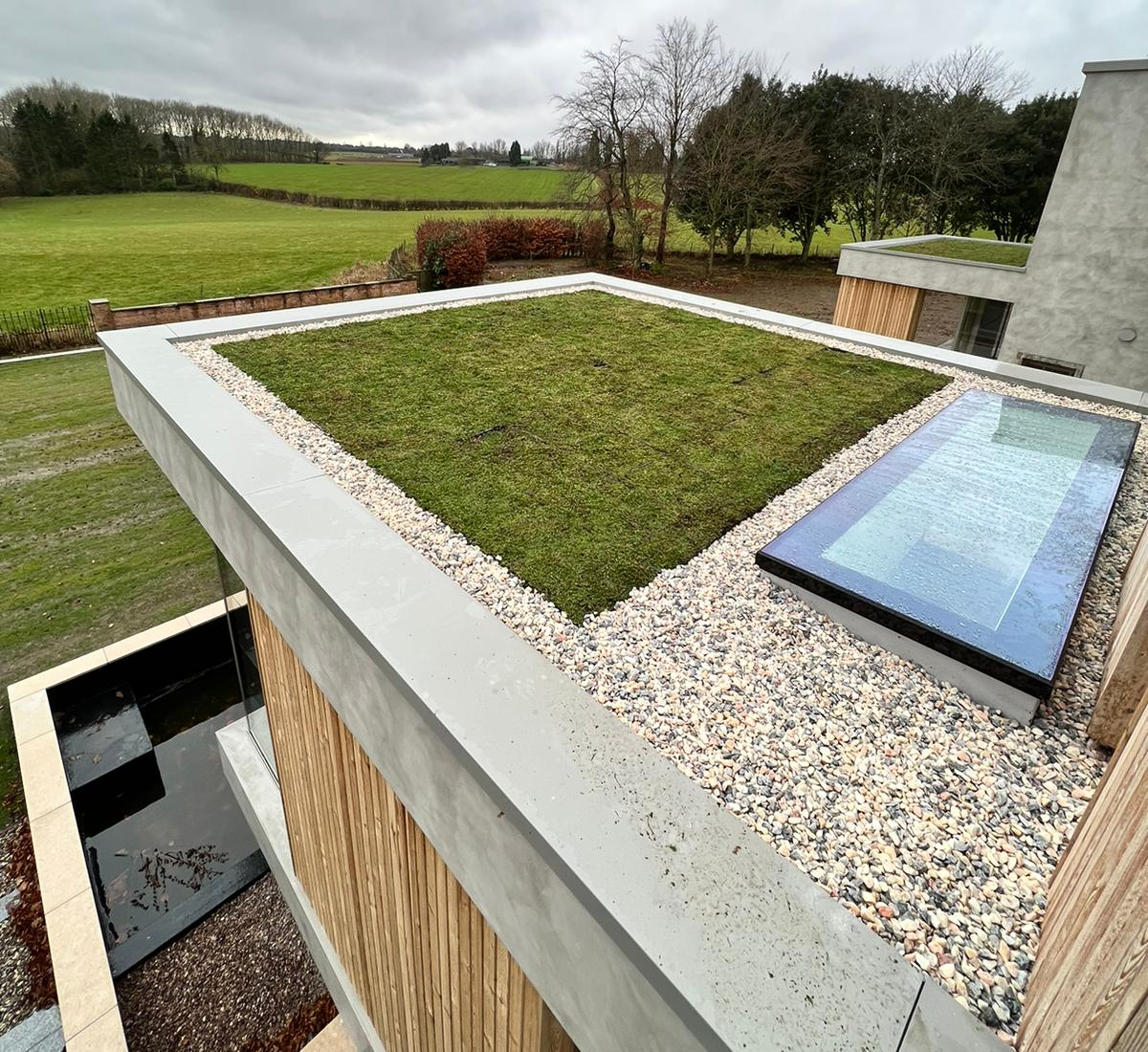 Rooftop with green lawn, gravel border, and skylight overlooking a green field under a cloudy sky.