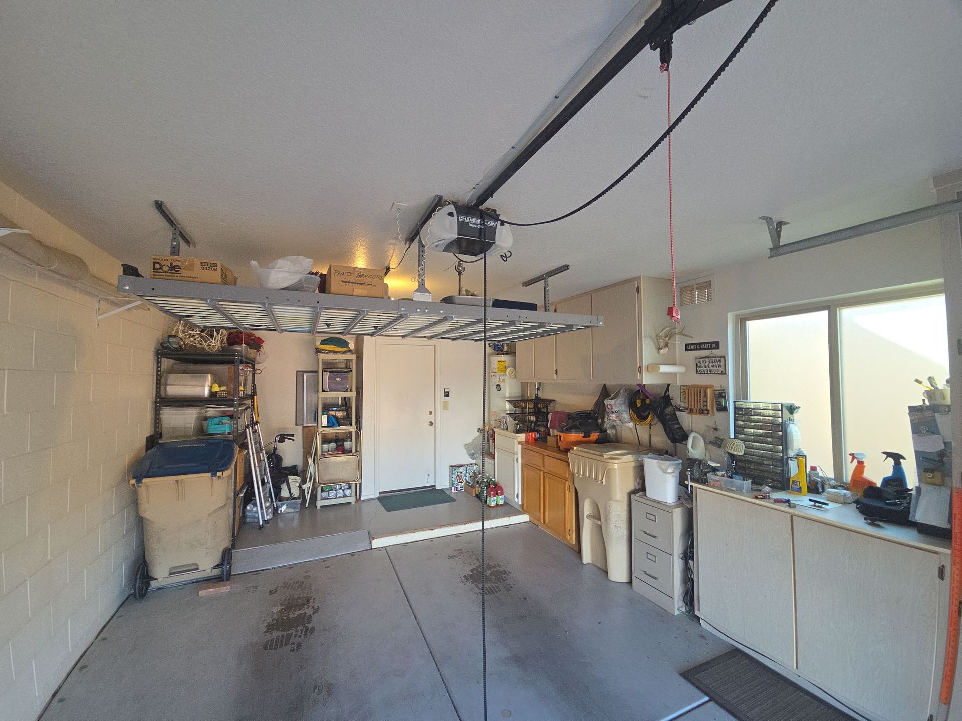 A cluttered garage with shelves, tools, cabinets, and a workbench under an open garage door.