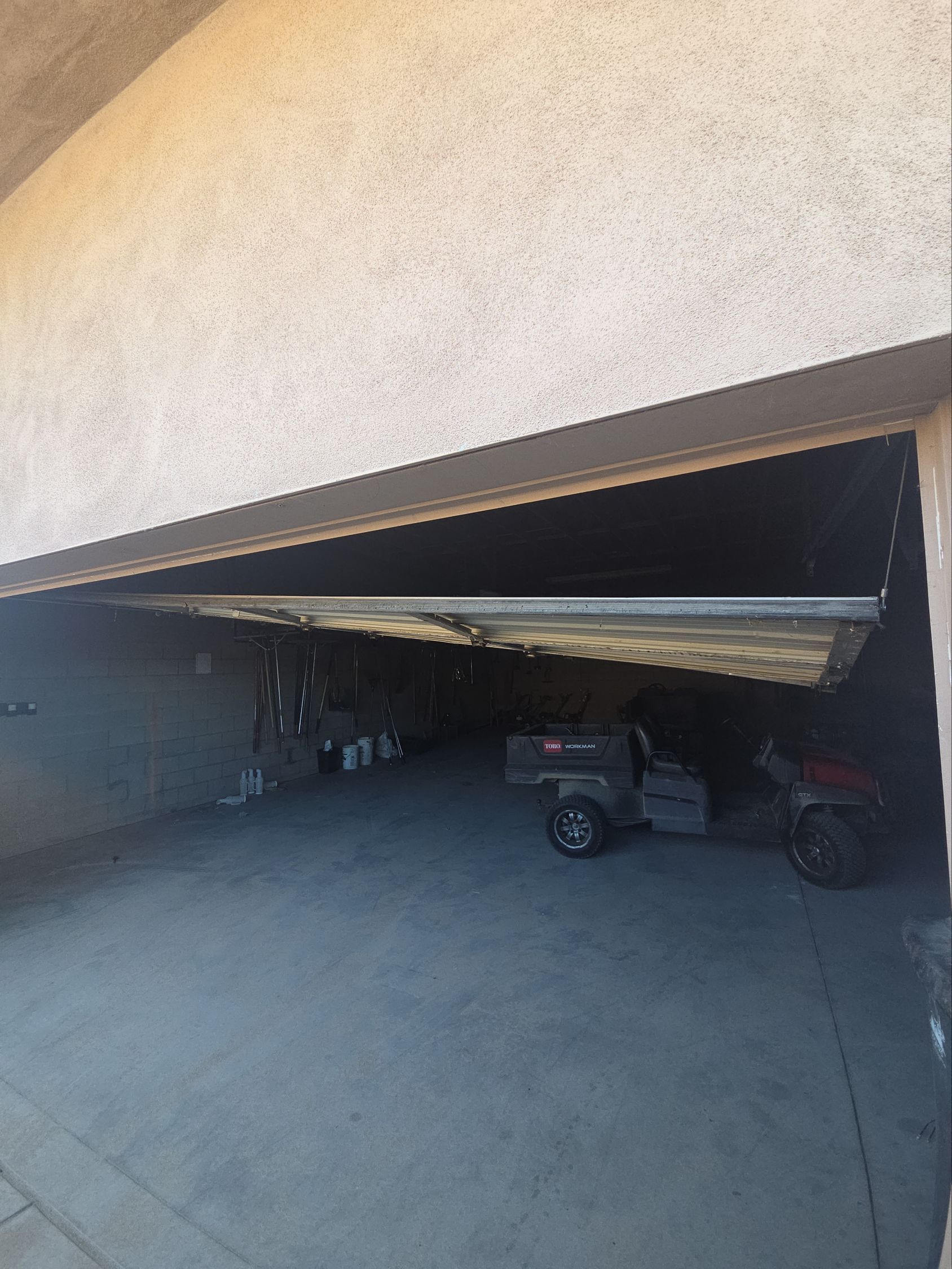 Partially open garage with a tilted overhead door, revealing a shaded interior and parked small vehicles.