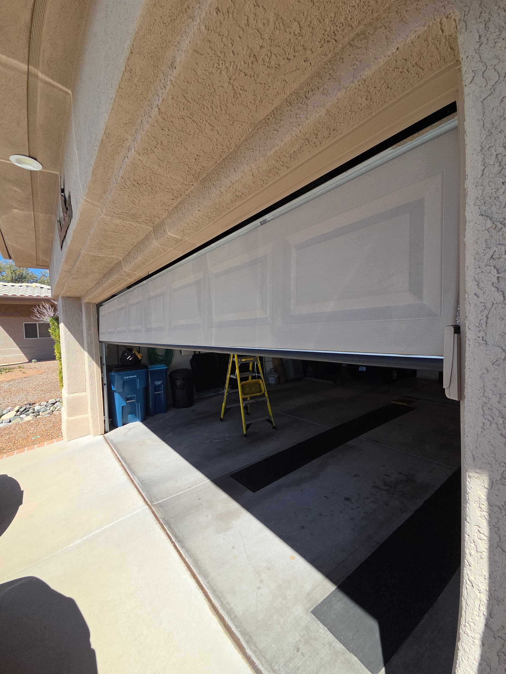 Partially open white garage door with yellow ladder inside, viewed from driveway under stucco overhang