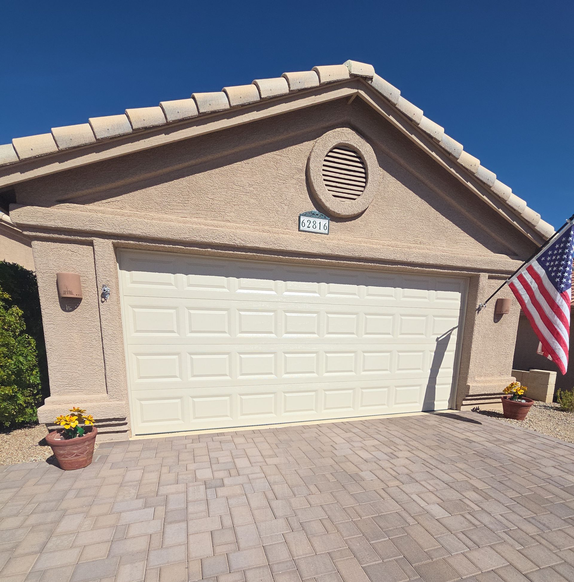 Stucco house front with white garage door, American flag, and potted flowers under a clear blue sky