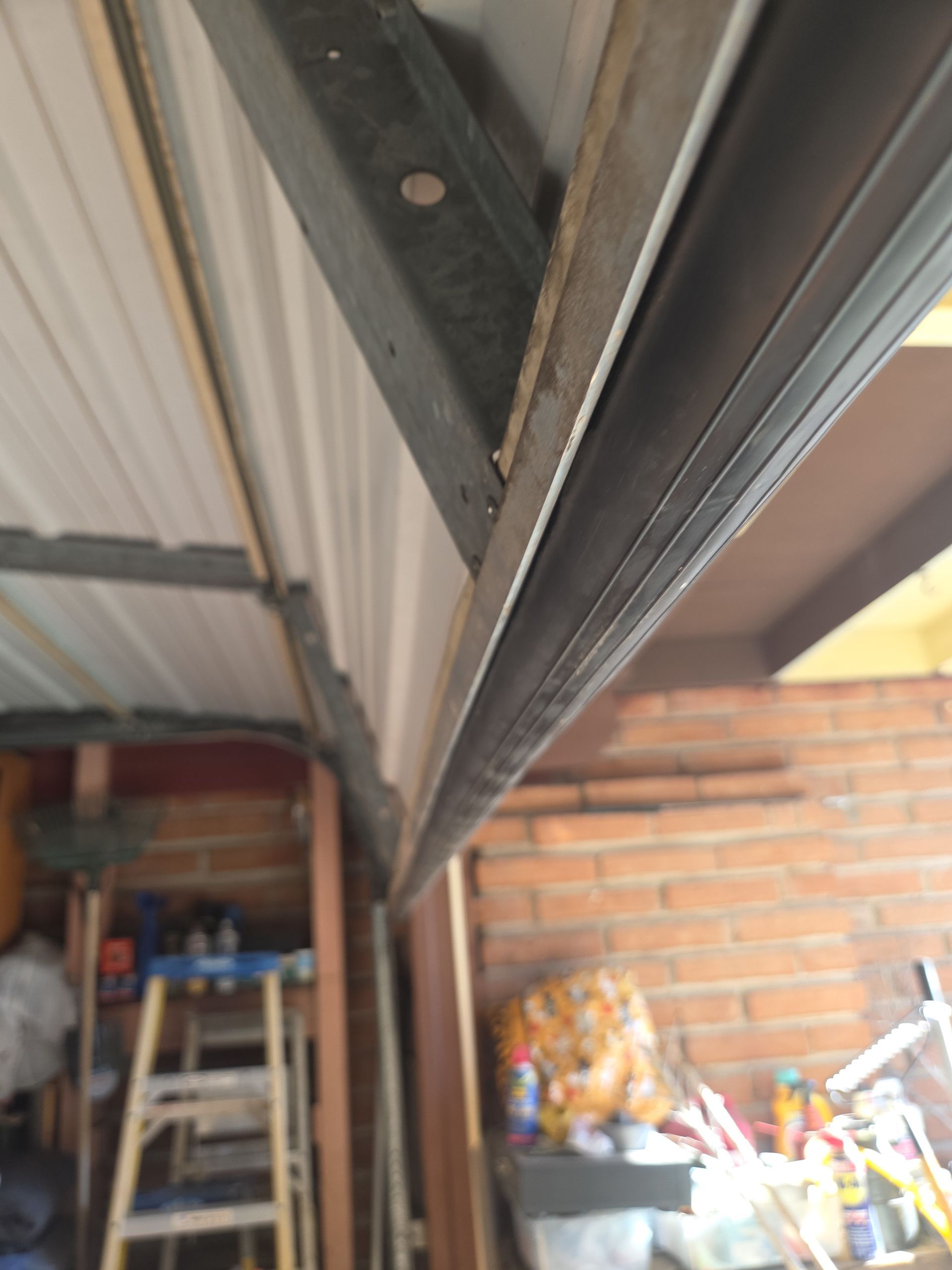 Close-up of a metal roof seam under a corrugated carport, with a ladder and brick wall in the background.