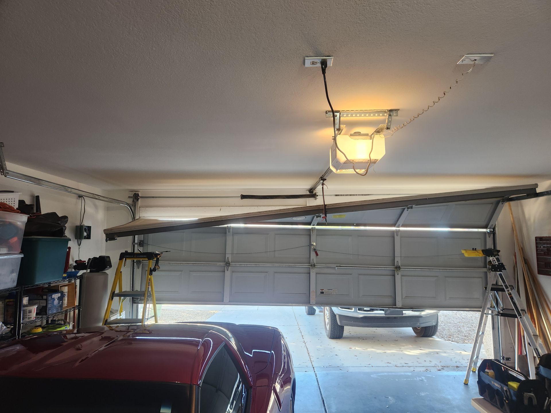 Garage interior with a partially open garage door, red car, and ladder beneath a ceiling light
