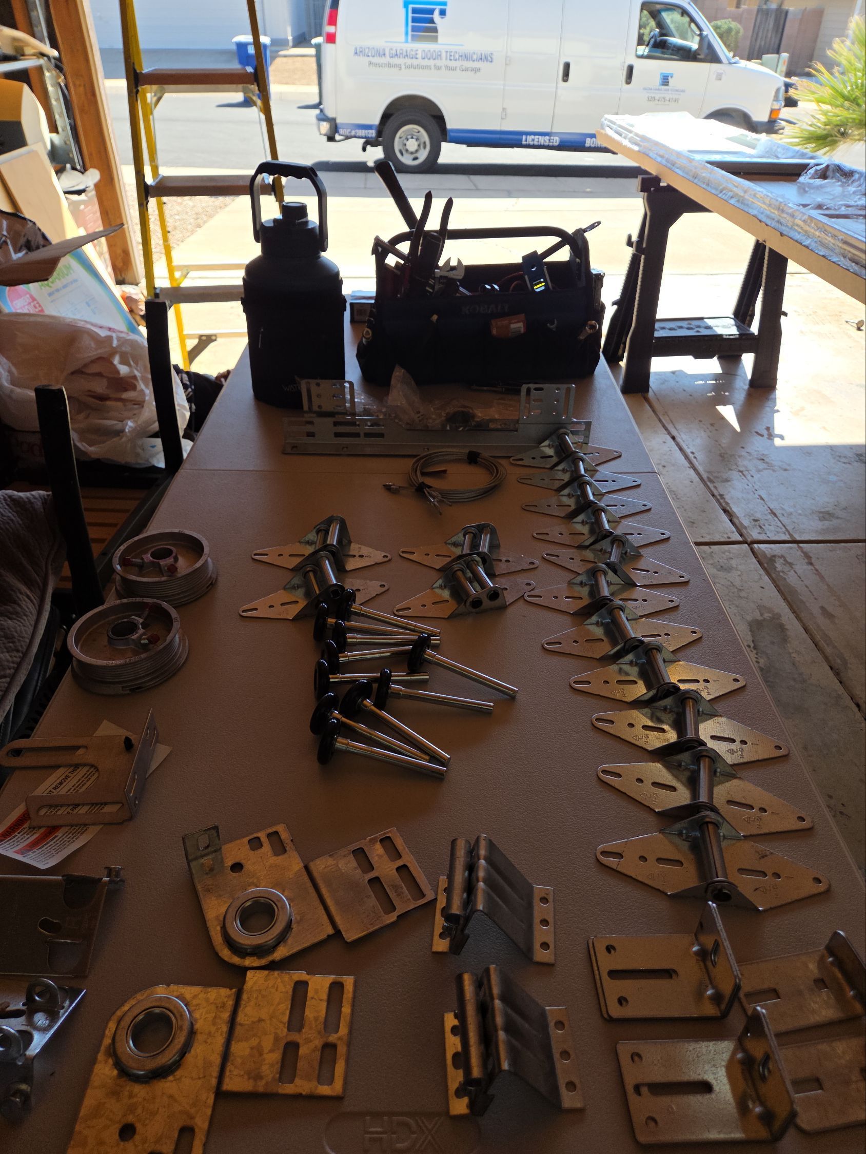 Tools and metal parts laid out on a workbench in a workshop, with a van outside in the background.