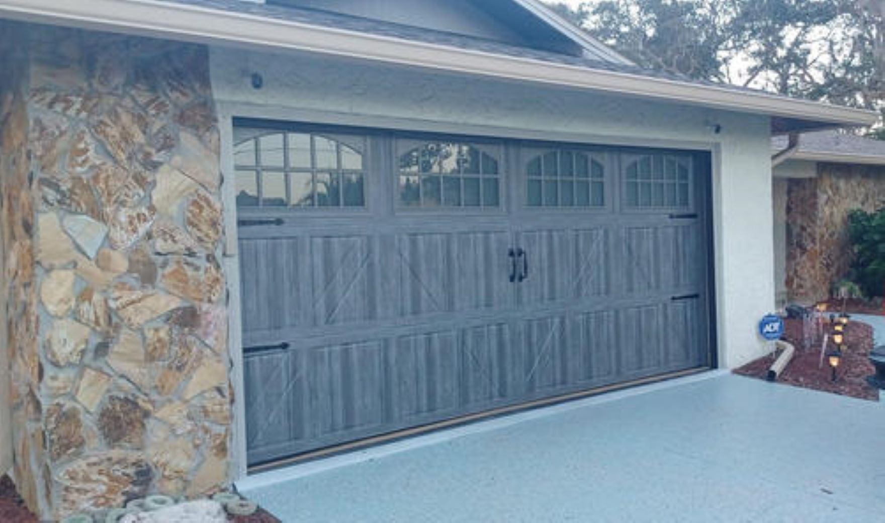 Gray double garage door with arched windows on a stone-front house and light blue driveway.
