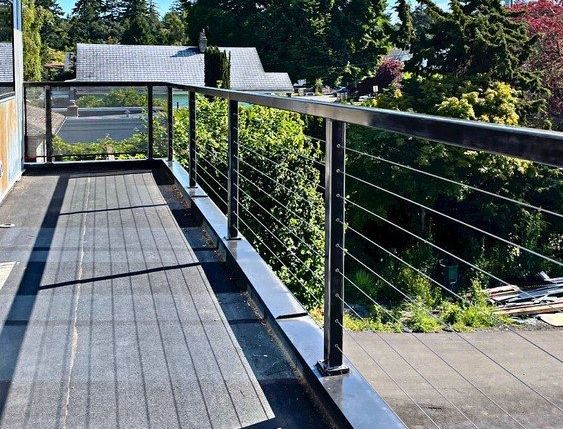 Outdoor balcony with dark decking, black railing, and green trees beyond, lit by bright sunlight