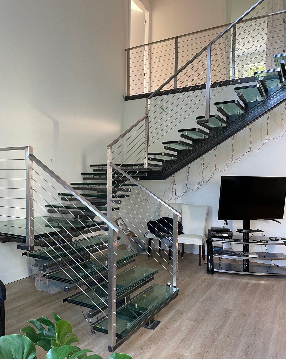 Modern living room with glass staircase, TV, white chair, and a potted plant on wood flooring