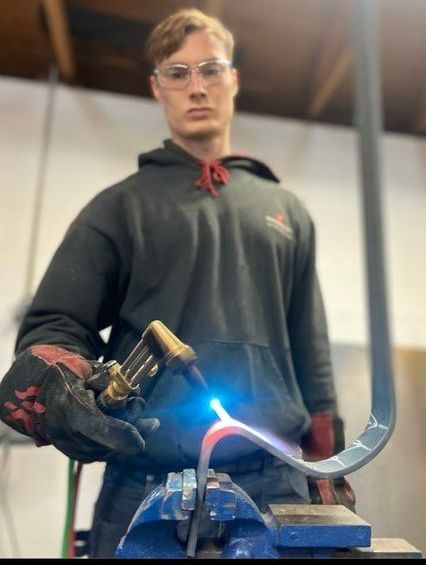 Person in safety glasses and gloves using a torch to heat a curved metal piece in a workshop.