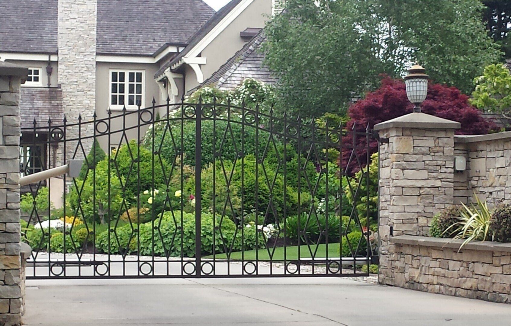 Black wrought-iron driveway gate in front of a landscaped stone house entrance