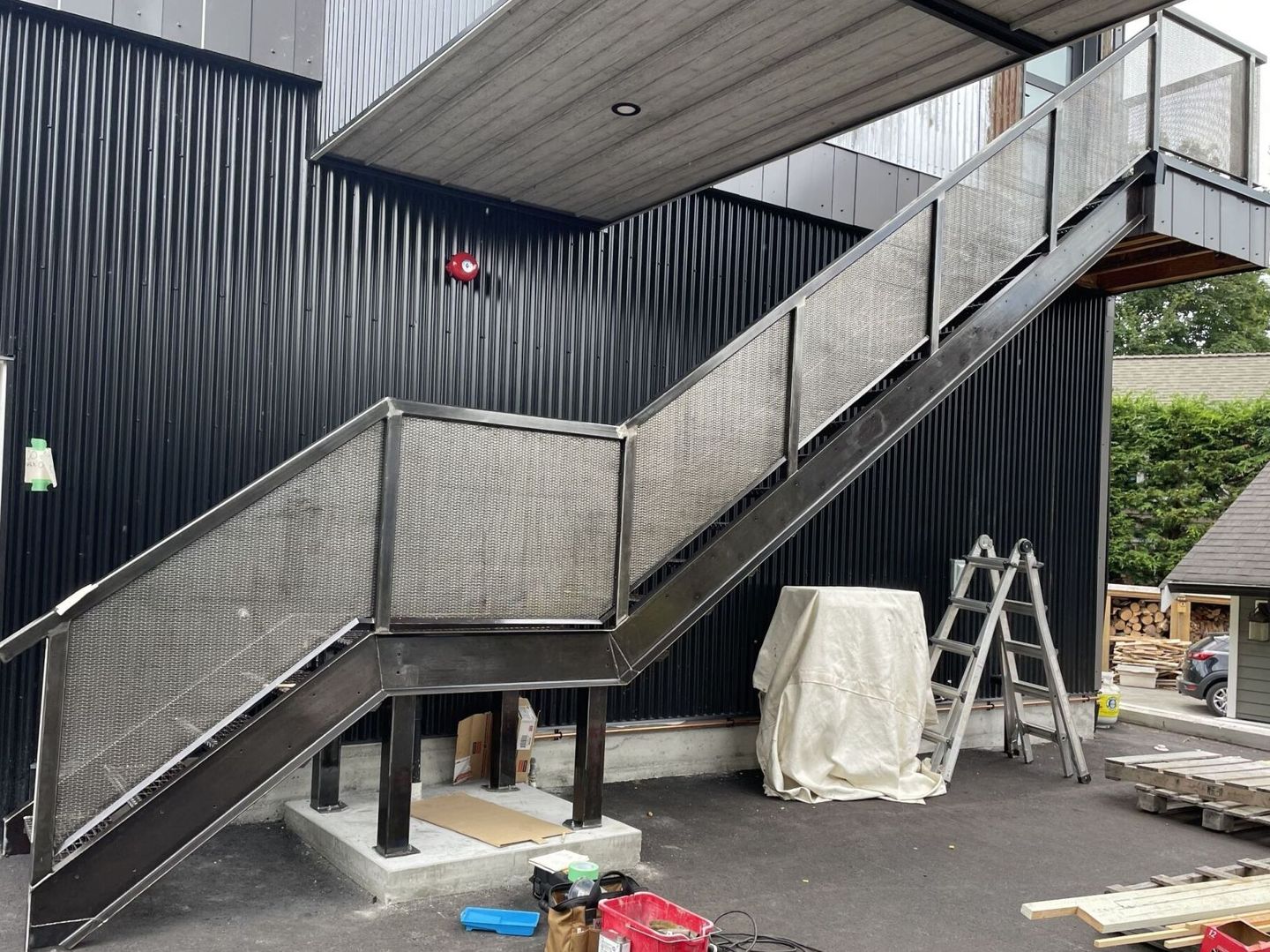 Metal staircase with mesh railings outside an industrial building, with ladders and covered items below.