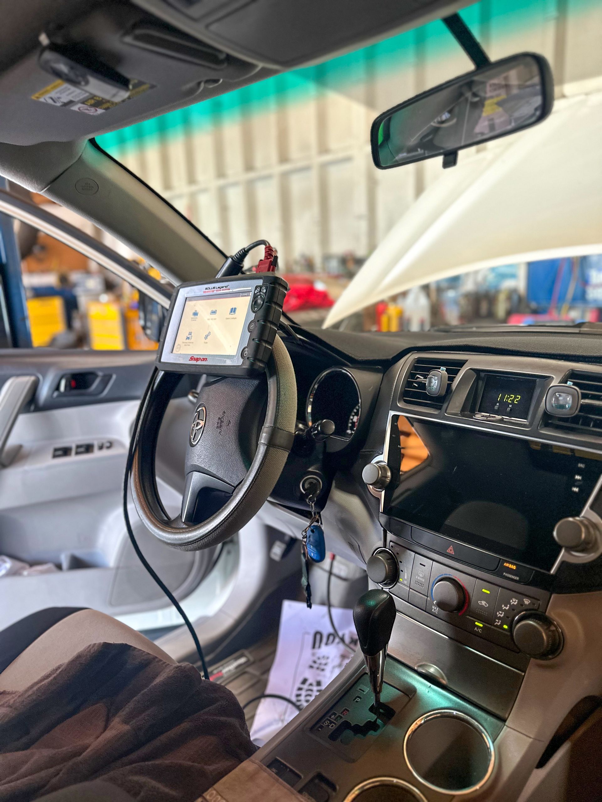 Interior car diagnostic setup: tablet attached to steering wheel, open hood in shop, papers & tools visible. | Jim Brangham Auto Repair