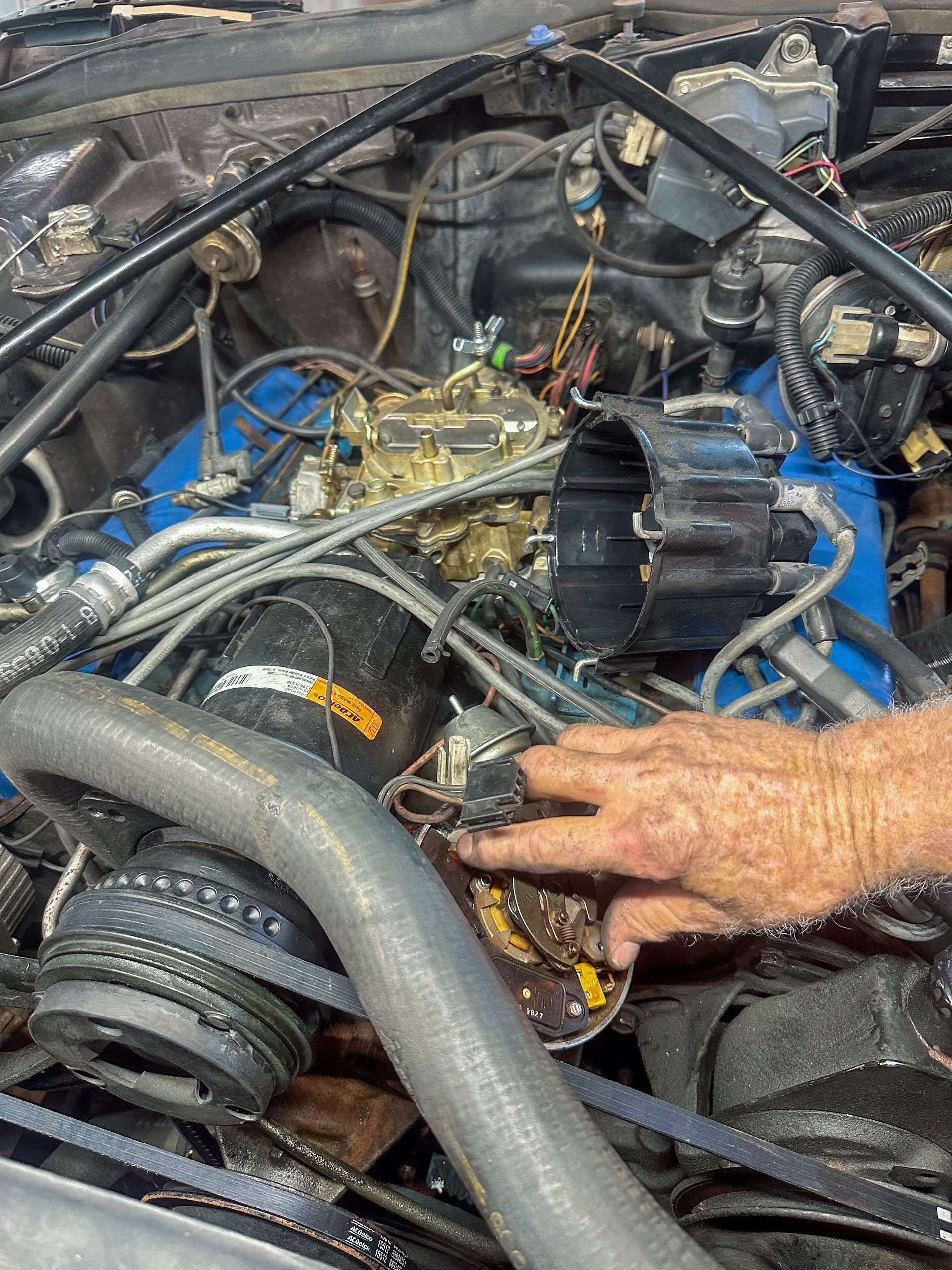 A person's hand points to the distributor of a blue engine in an older car. | Jim Brangham Auto Repair