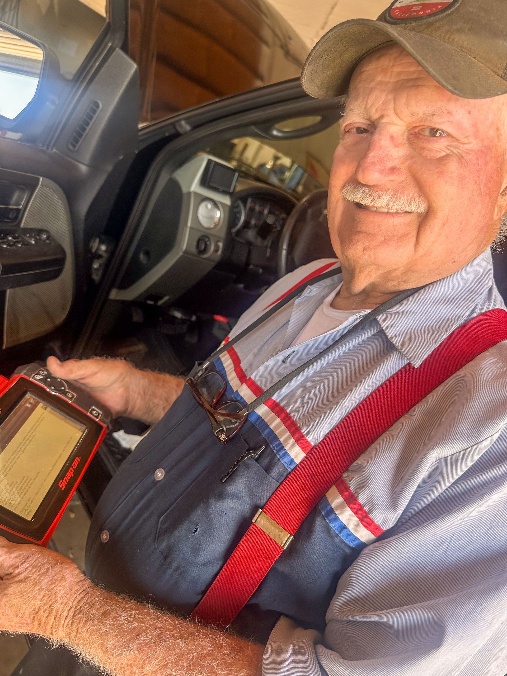 Elderly man in a work shirt, suspenders, and cap, holding a diagnostic tool inside a car, smiling. | Jim Brangham Auto Repair