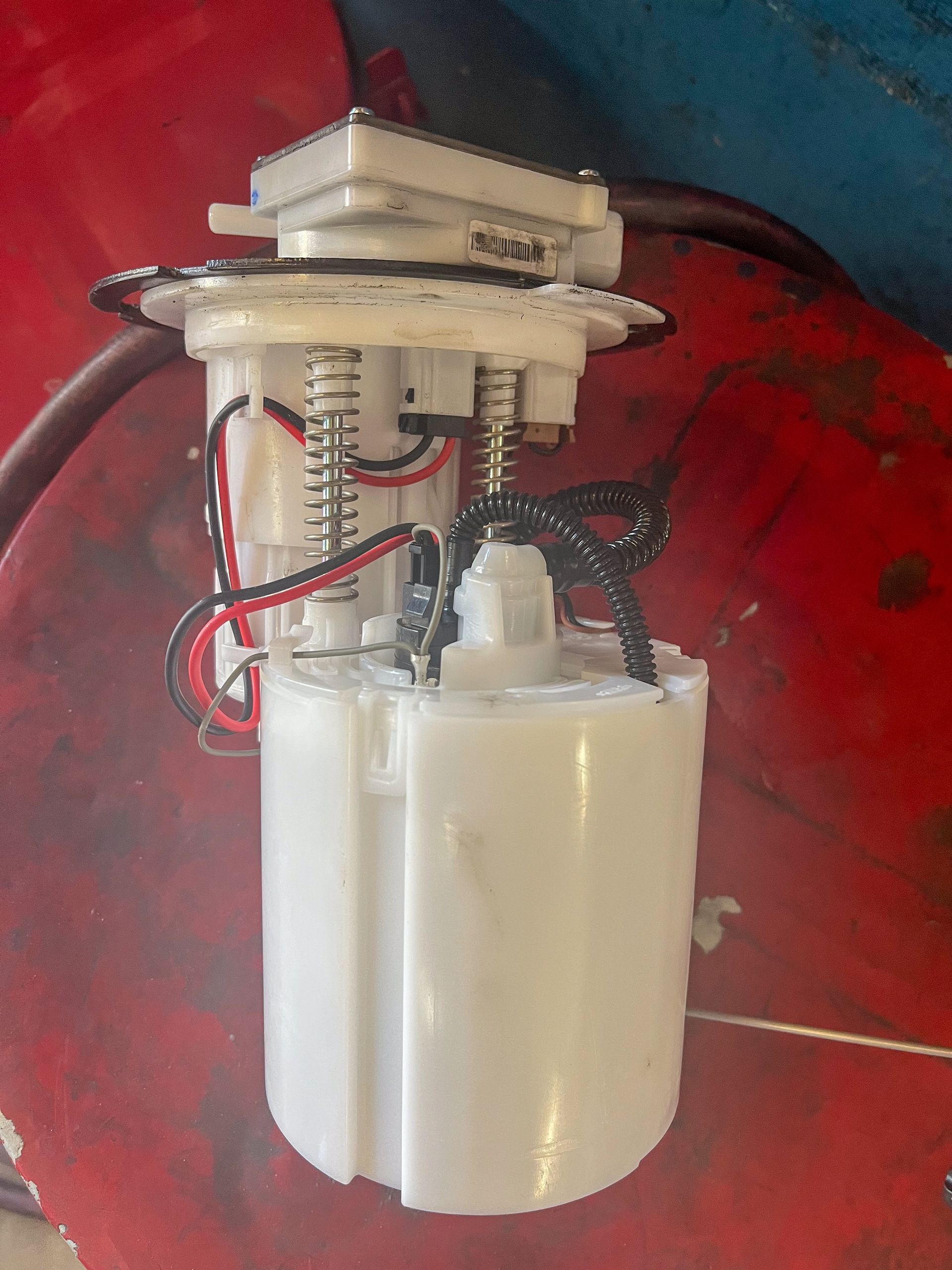 White fuel pump assembly on a red surface, with wires and a spring visible. | Jim Brangham Auto Repair