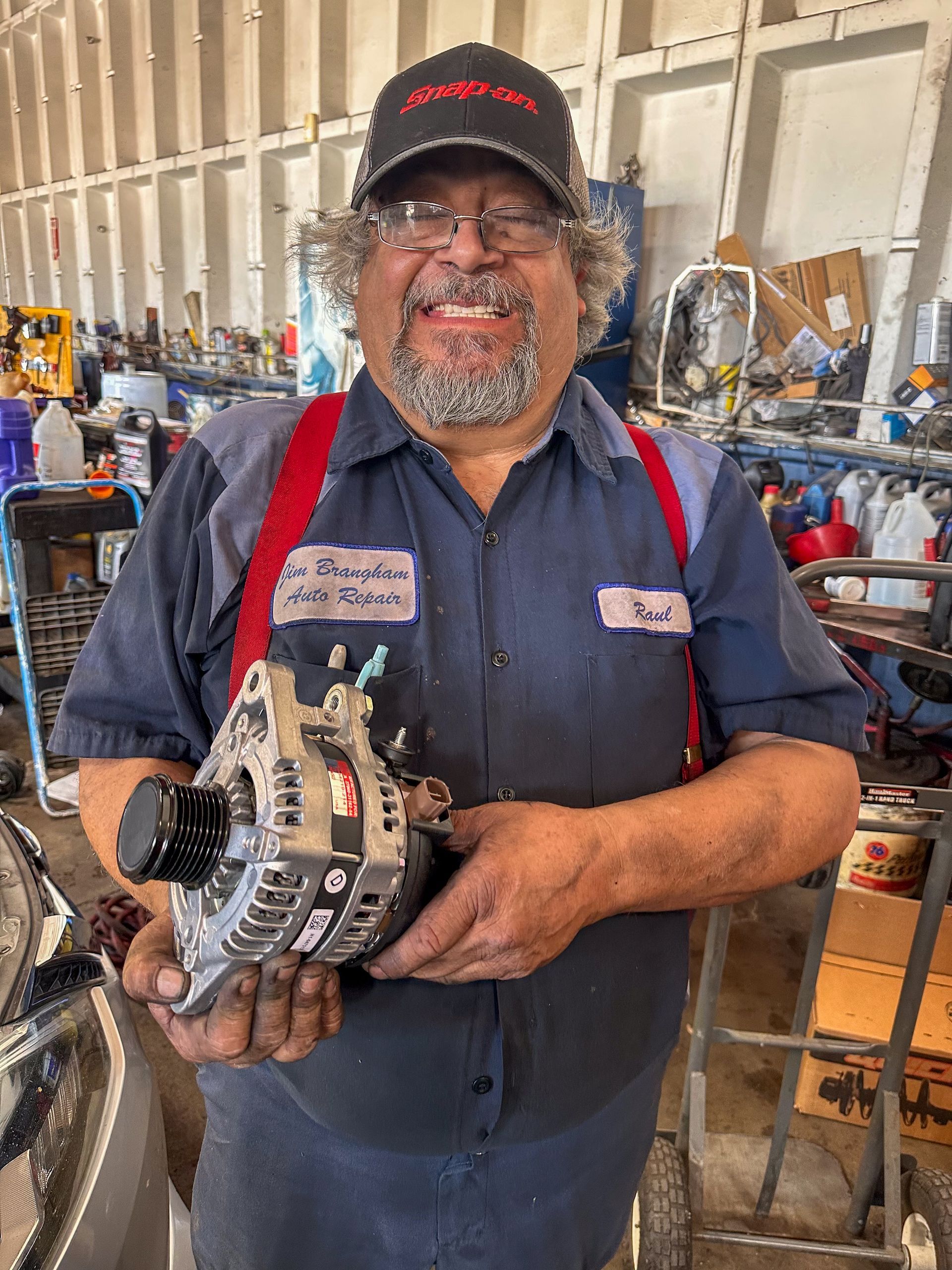 Smiling mechanic holding an alternator in a garage, wearing a hat and overalls. | Jim Brangham Auto Repair