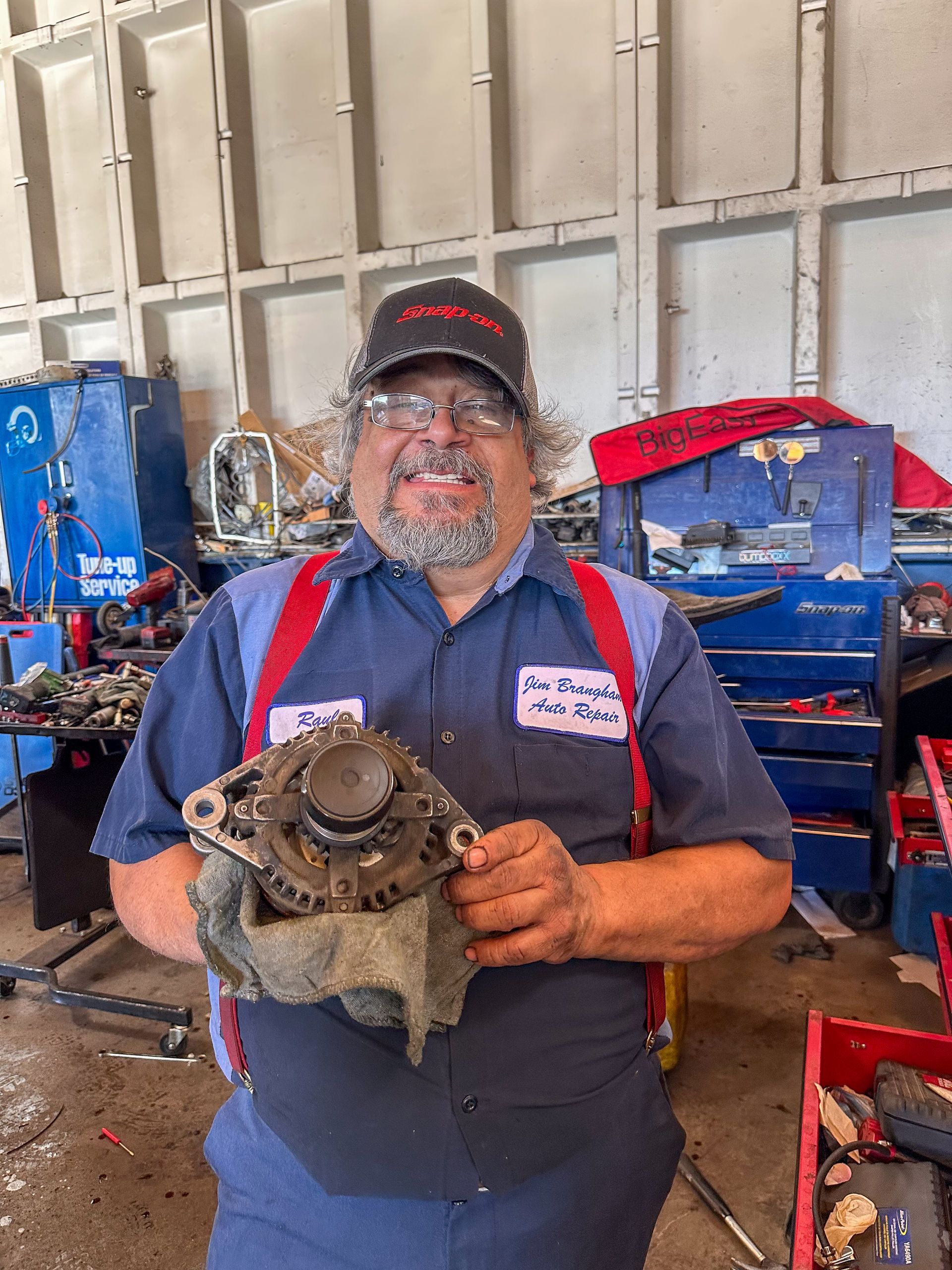 Mechanic holds up alternator. In auto shop, he smiles. He wears a hat, glasses, and work clothes. | Jim Brangham Auto Repair
