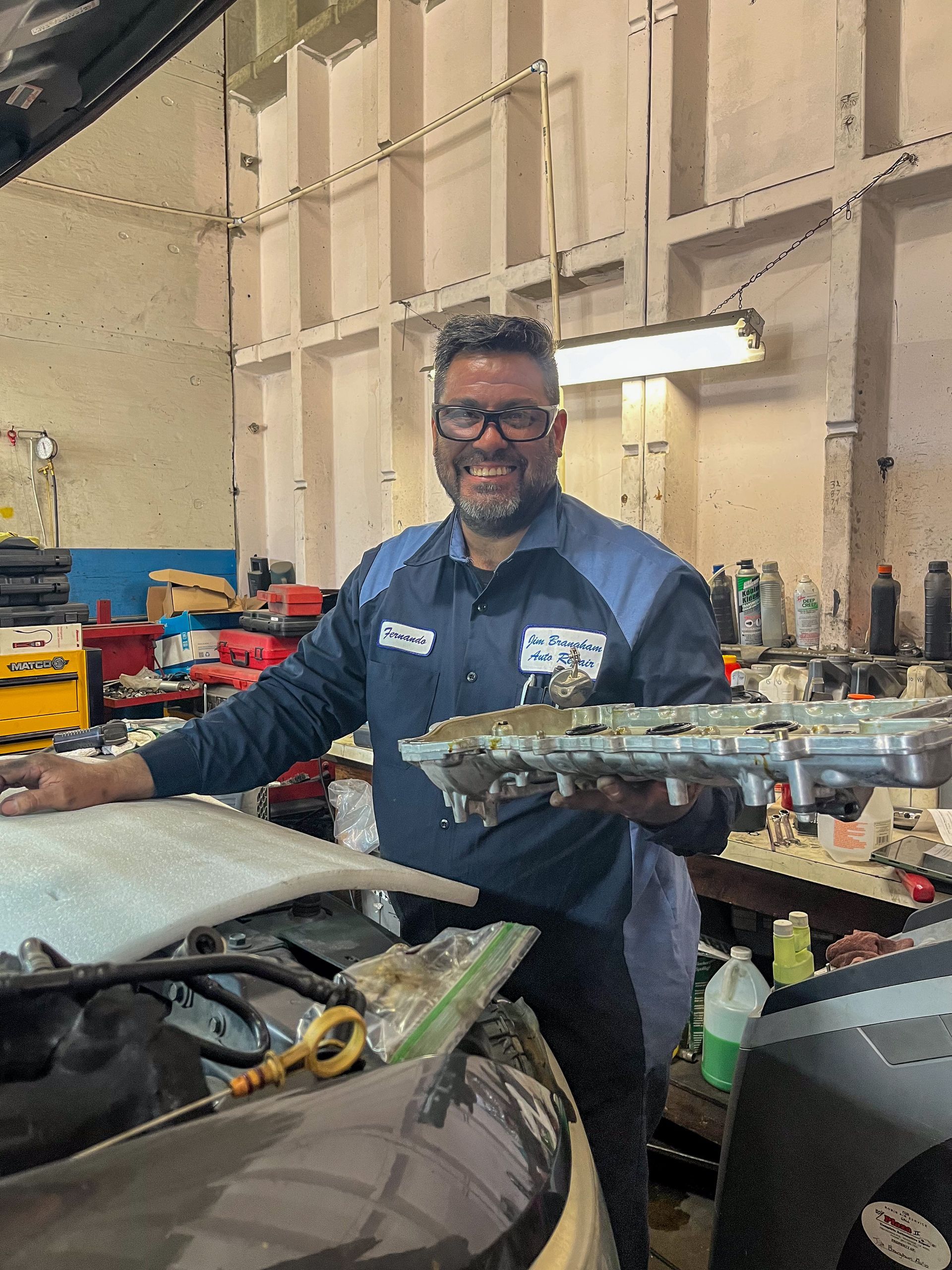 Mechanic holds car part in auto shop. Man in blue shirt smiles, standing near open car hood. | Jim Brangham Auto Repair