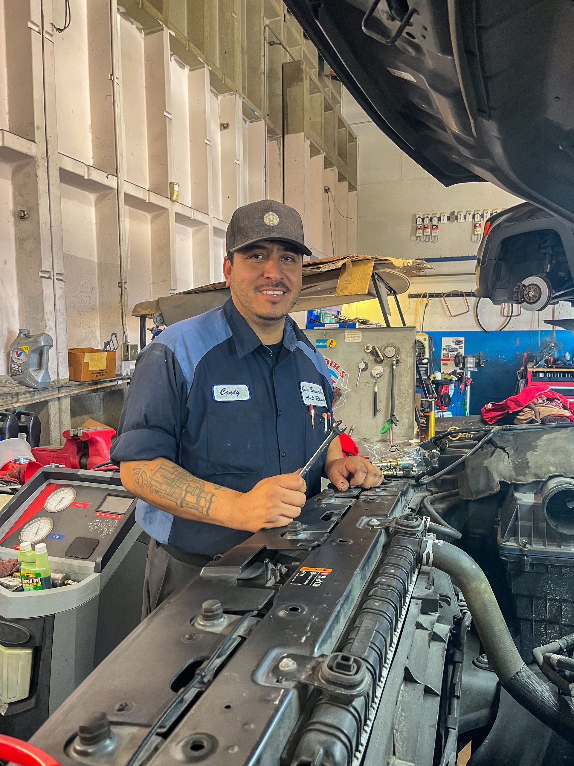 Mechanic in a shop, working on a car engine, smiling at the camera. Wearing a cap and uniform. | Jim Brangham Auto Repair