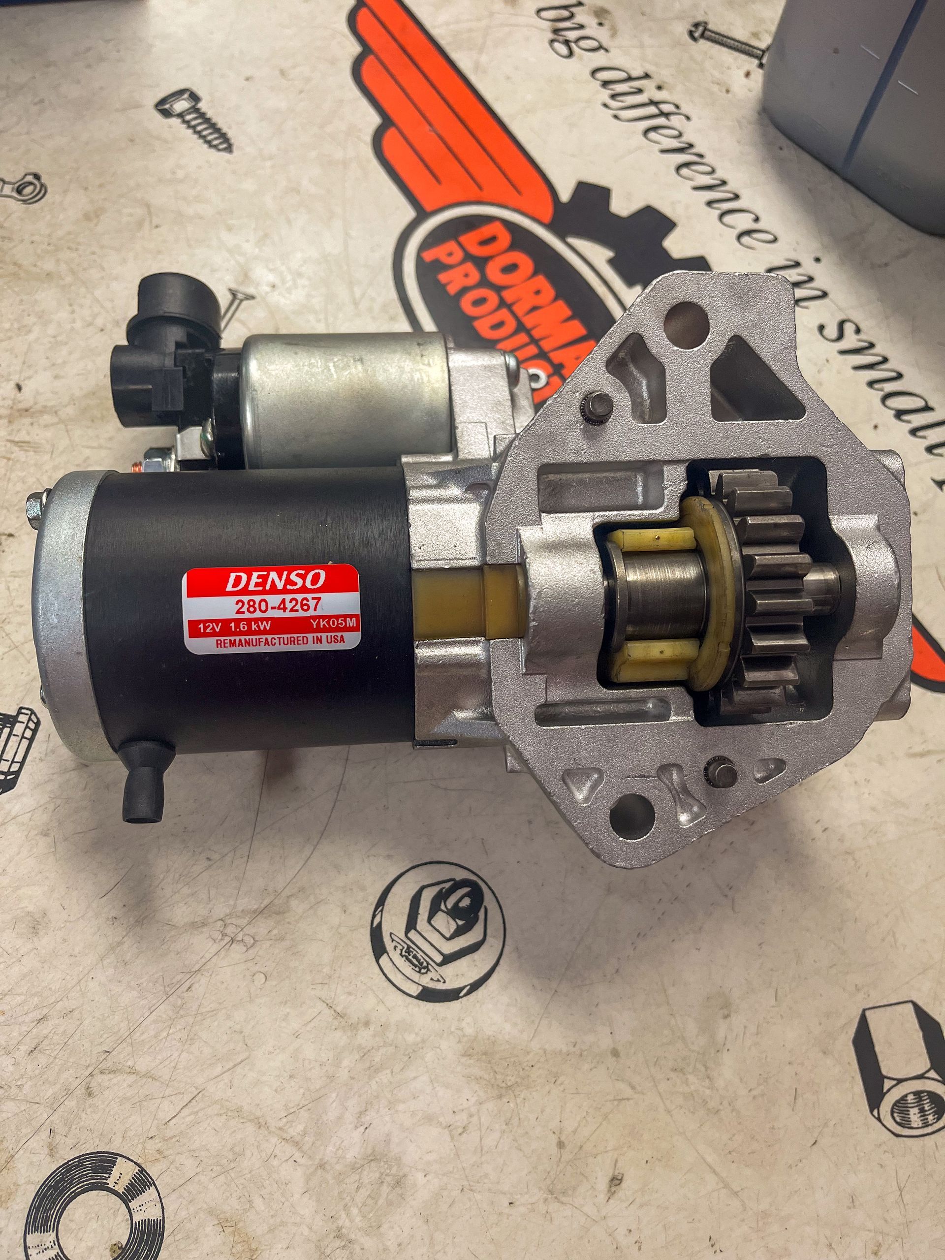 Denso starter motor, black and silver, with exposed gear, on a work surface with a Dorman Products logo. | Jim Brangham Auto Repair