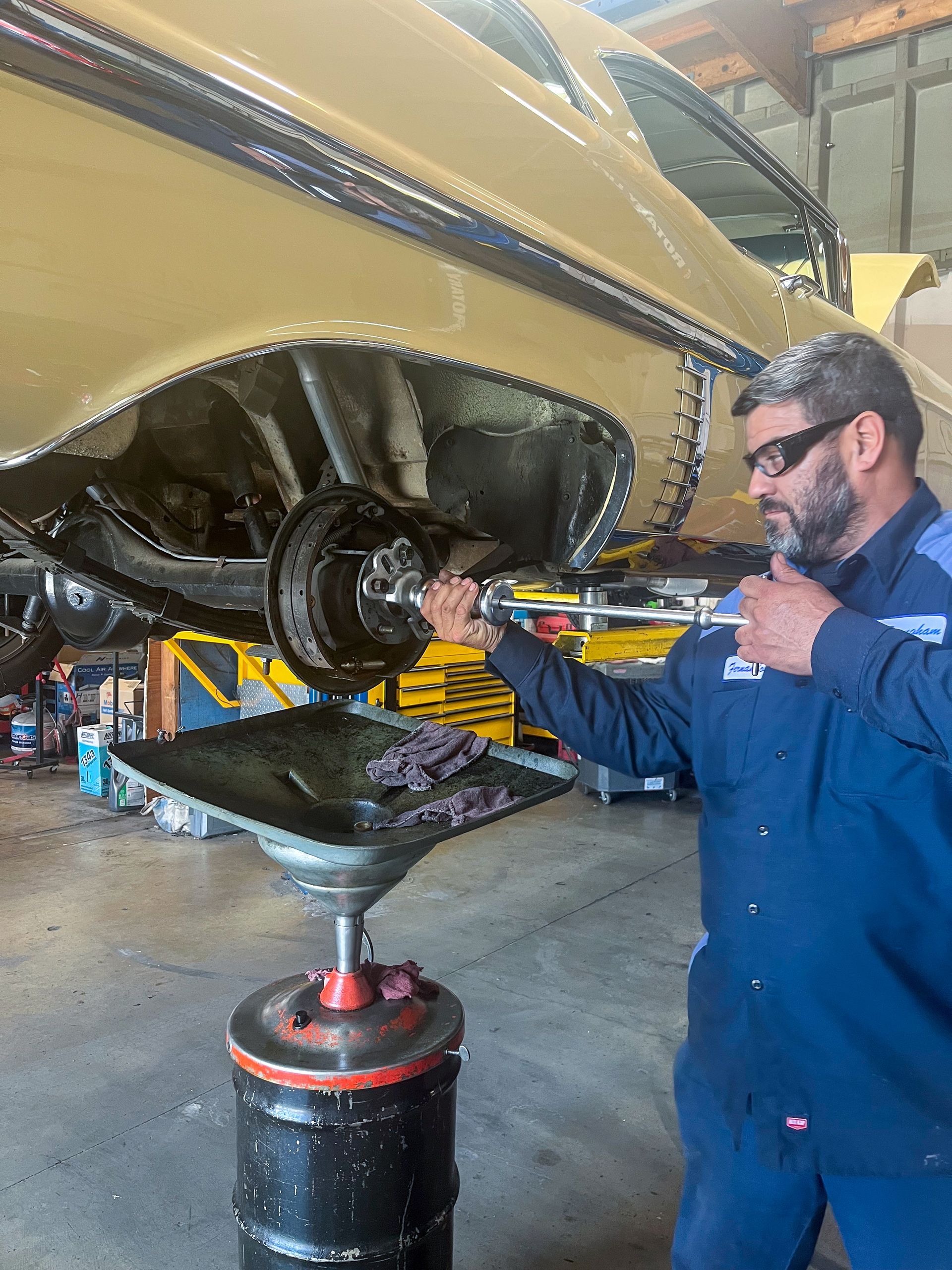Mechanic working on car’s undercarriage. He’s wearing blue overalls and looking at the rear axle, with a oil drain pan in the foreground. | Jim Brangham Auto Repair