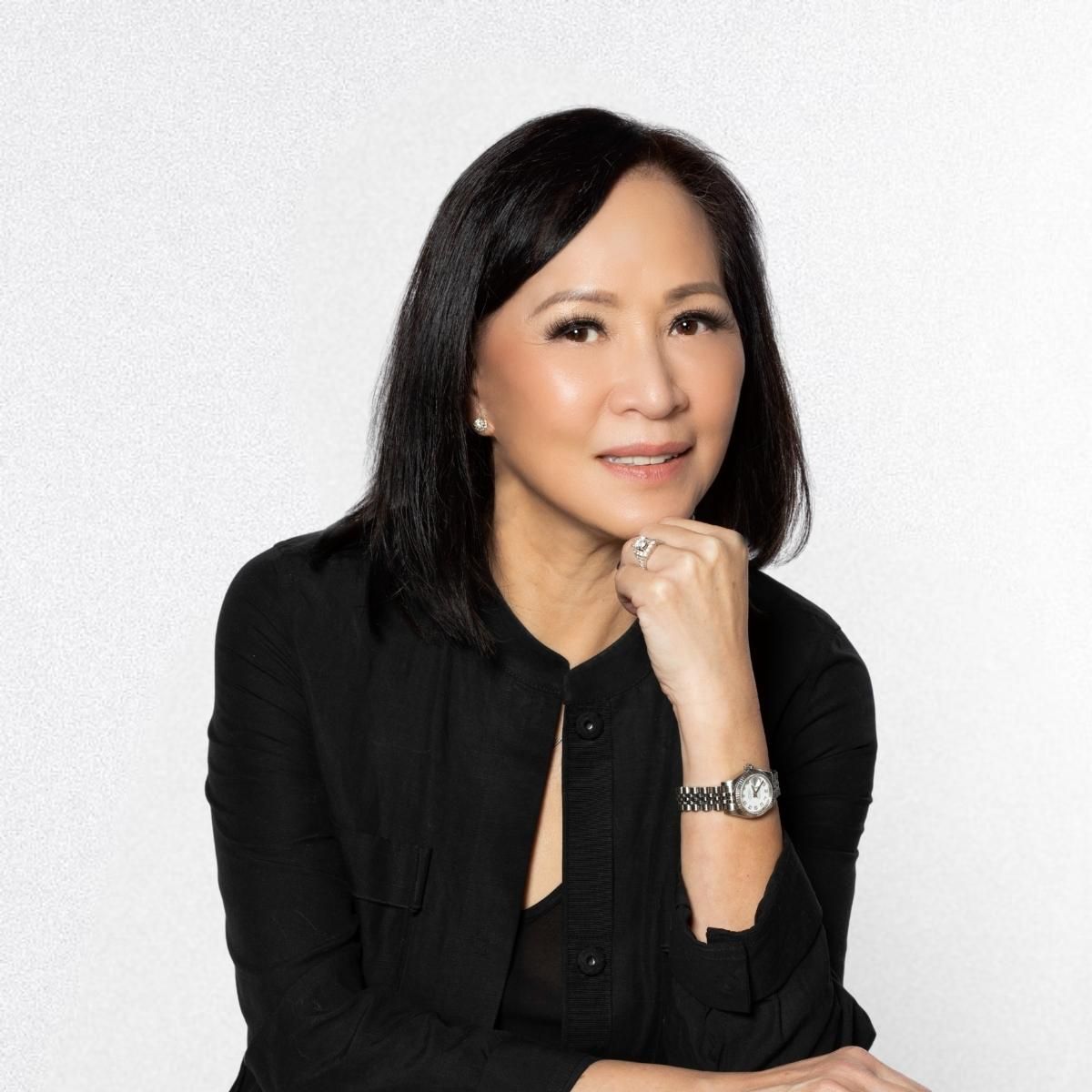 A woman wearing a black shirt and a watch is sitting with her hand on her chin.